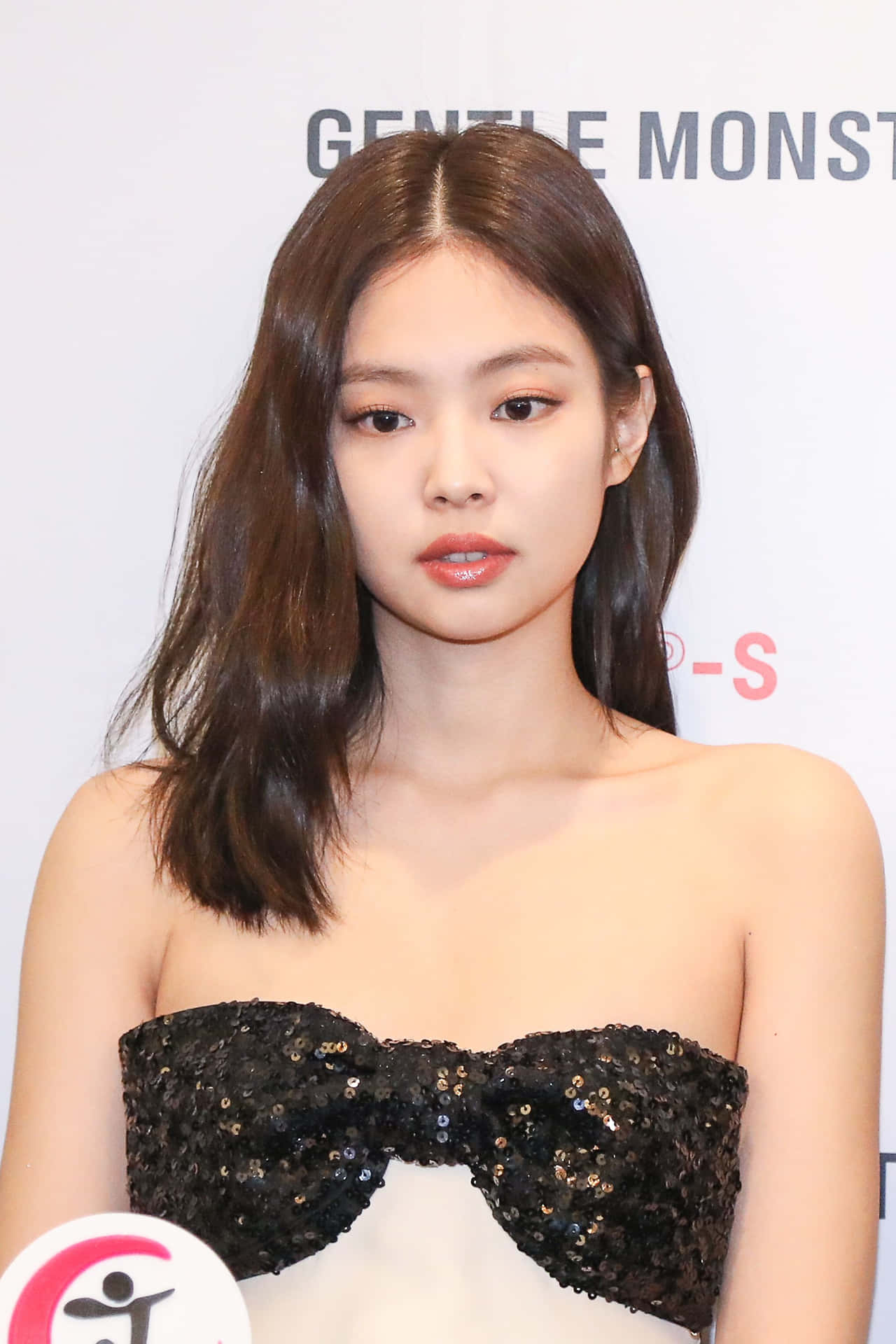 Jennie BLACKPINK 紅毯亮片禮服快照 壁紙 — 一位留著及肩棕色長髮的女性，在白色品牌背景前穿著黑色閃片露肩上衣。