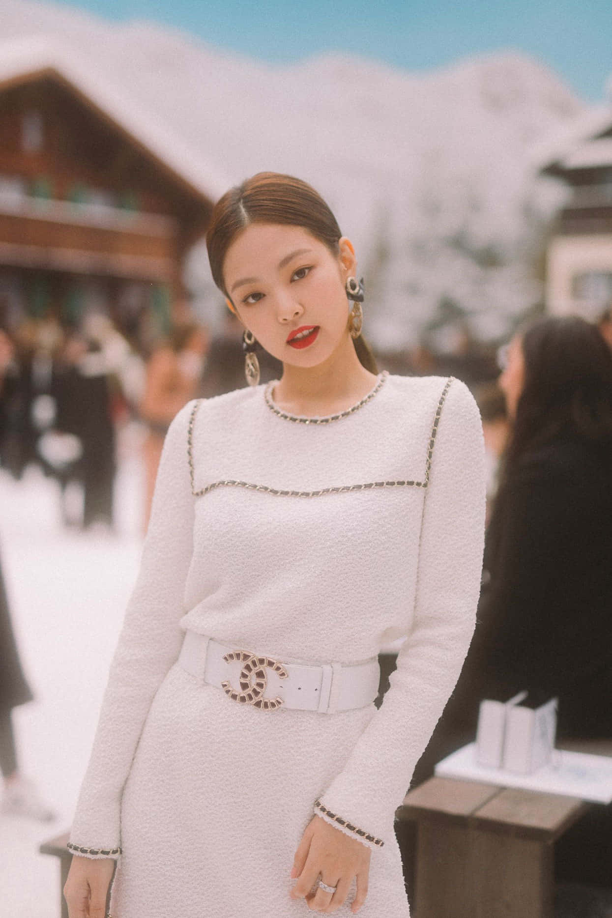 Jennie BLACKPINK 穿香奈兒禮服照 壁紙 — 一位身穿白色粗花呢 Chanel 連衣裙、搭配標誌腰帶的女性，自信地站在白雪覆蓋的山村環境中。