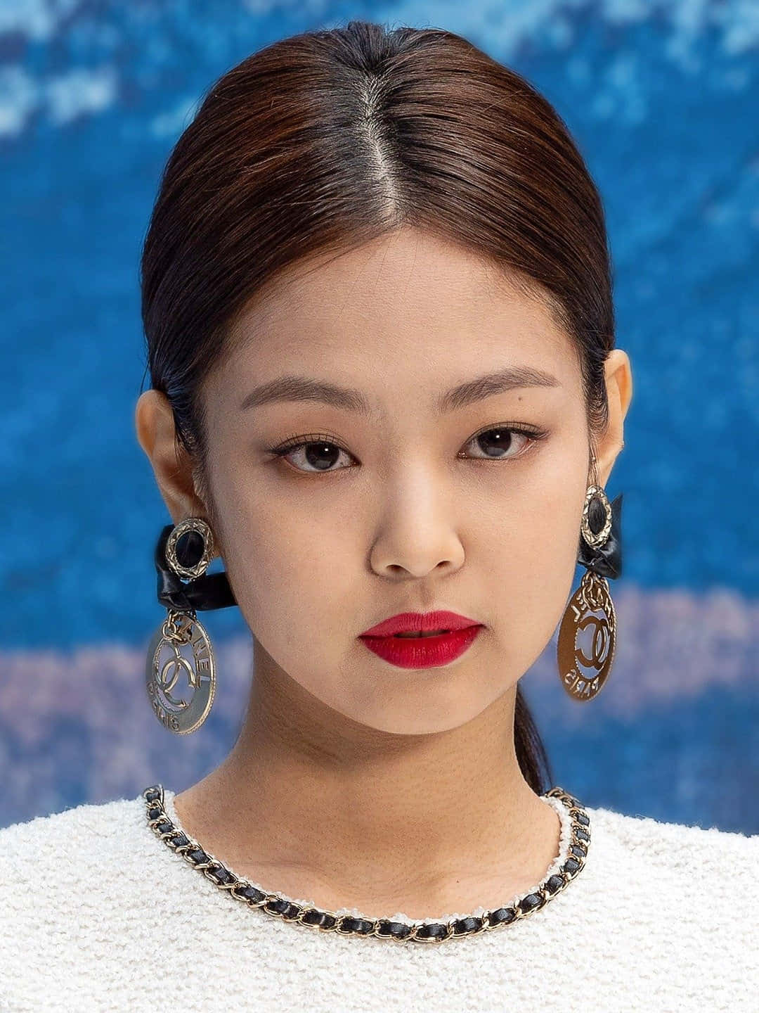 Jennie Kim 紅唇耳環美照 壁紙 — 一位擁有光滑黑髮和大膽紅唇的女性，在帶有紋理的藍色背景前佩戴華麗的香奈兒耳環。