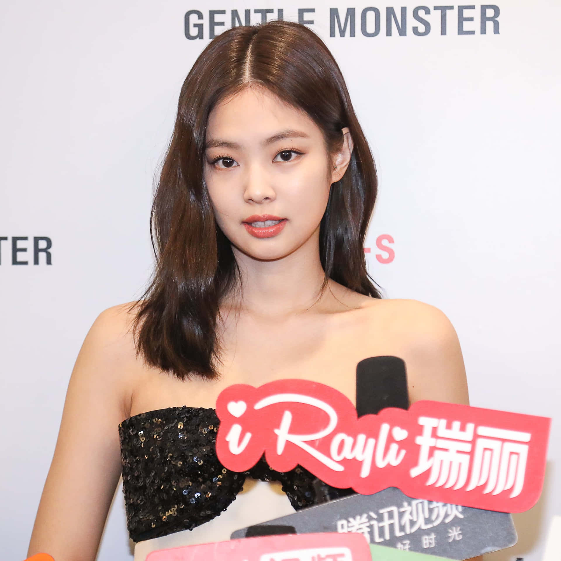 Jennie Kim 紅毯快照 壁紙 — 一位留著及肩棕色長髮的女性，在設有咪高峰和「GENTLE MONSTER」背景的記者會上，穿著黑色閃片無肩帶上衣。