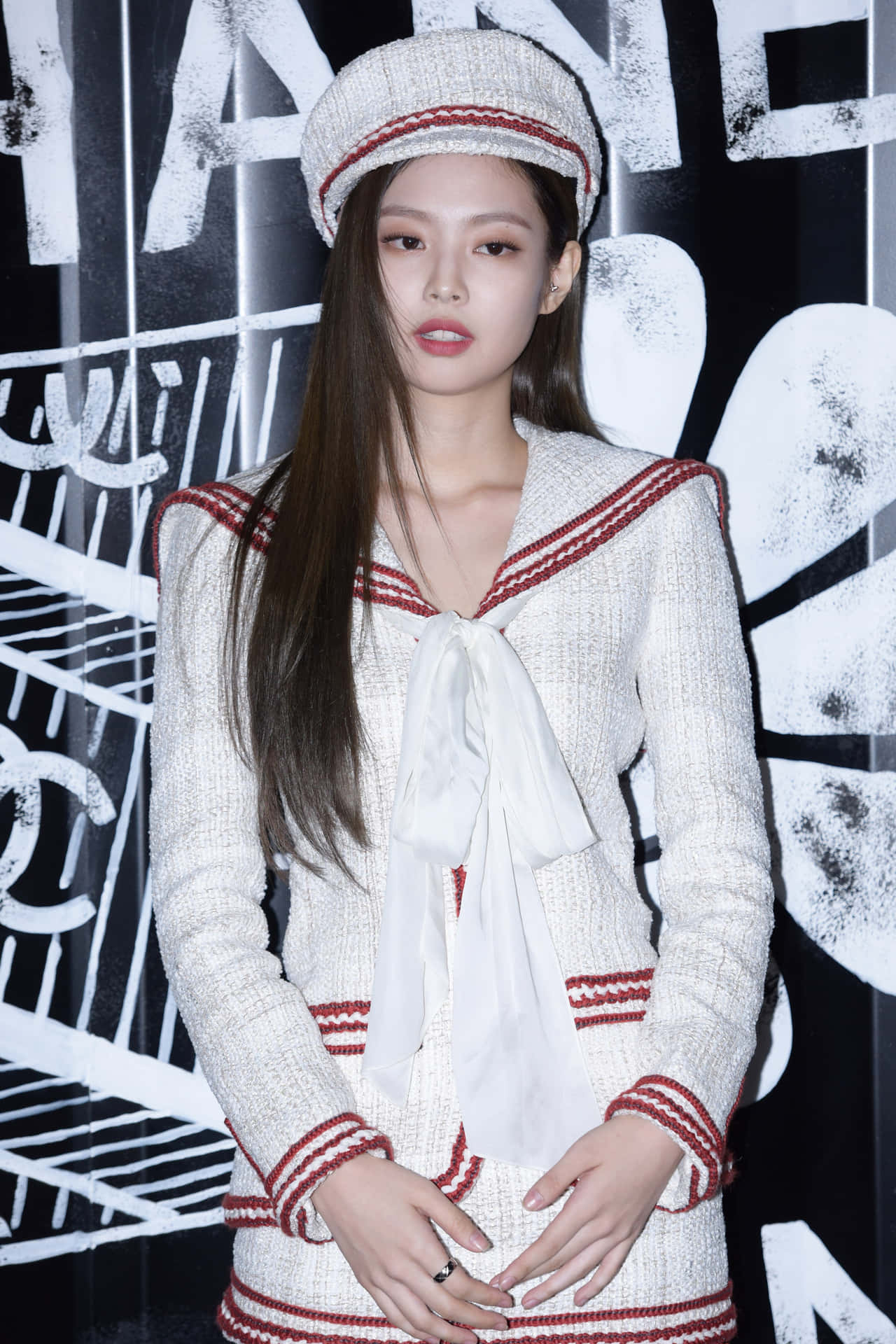 Jennie Kim 紅白禮服紅毯照 壁紙 — 一位優雅的女士身穿白色粗花呢水手服，配有紅色飾邊和相配的帽子，站在大膽的黑白 Chanel 標誌背景前。