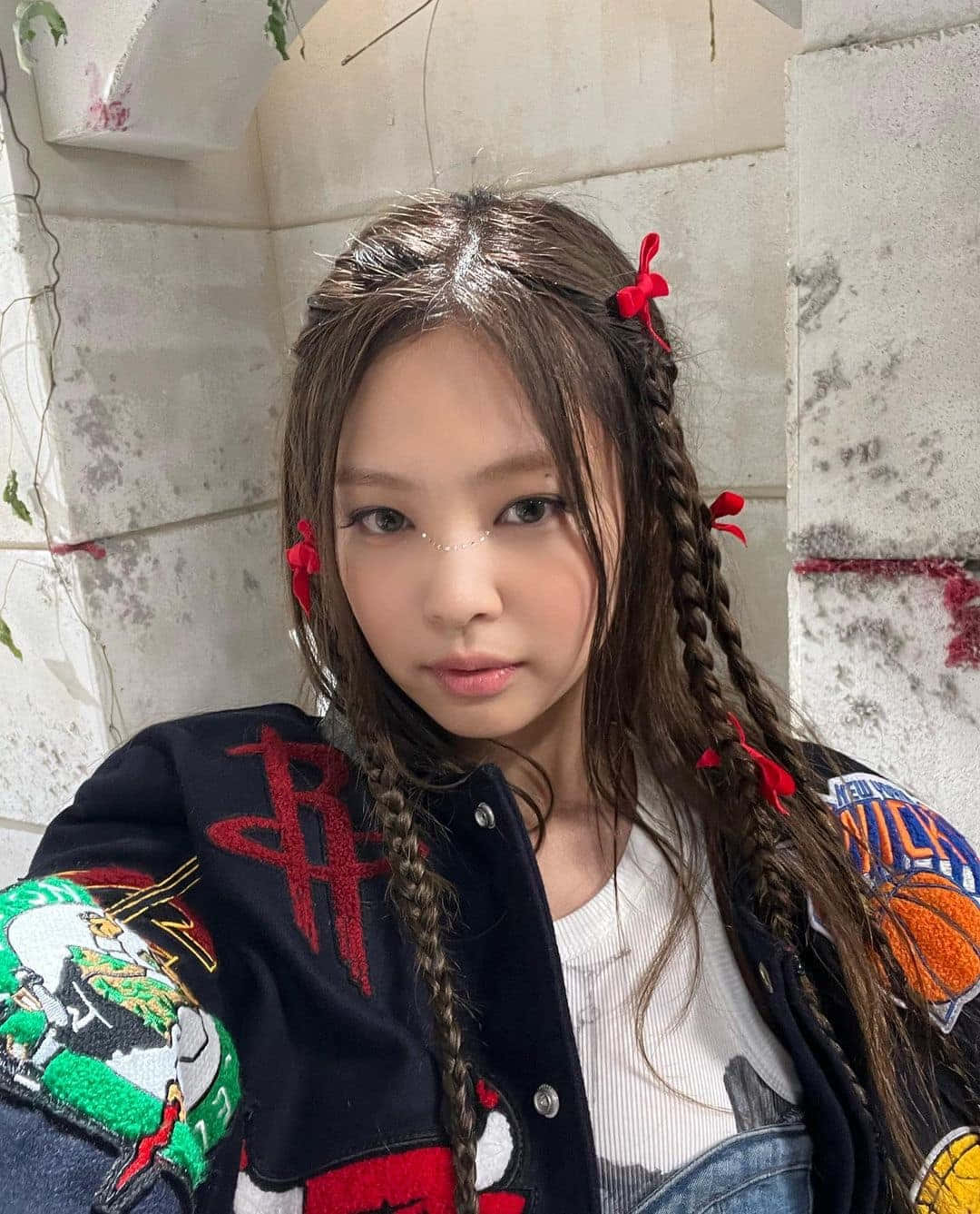 Jennie Kim 編髮配小絲帶造型照 壁紙 — 一位年輕女子，辮子上繫著紅色絲帶，身穿一件打滿補丁的夾克，靠著風化的石牆。