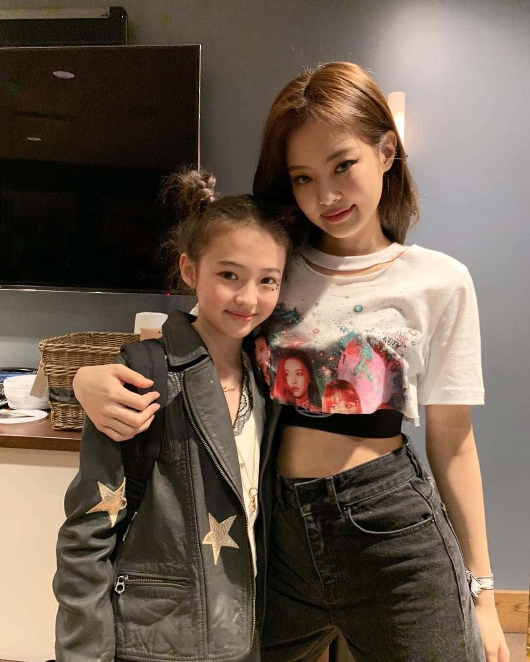Jennie Kim 與小女孩合照 壁紙 — 兩位女性在室內溫馨地擺姿勢，一位身穿星星外套，另一位身穿圖案T恤，背景有電視和柳條籃。