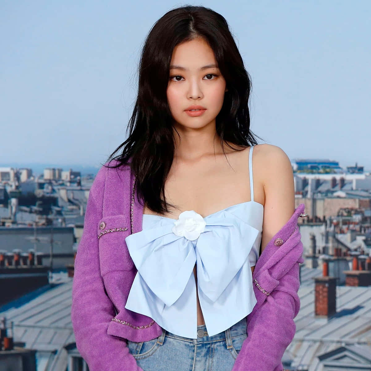 Jennie Kim 藍色上衣綁帶照 壁紙 — 一位身穿紫色外套和淺藍色蝴蝶結上衣的年輕女性自信地站在城市天際線背景下。