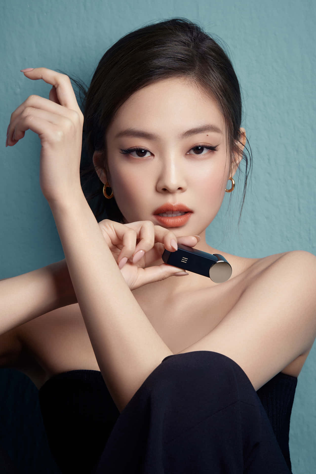 Jennie Kim 黑色禮服性感照 壁紙 — 一位深色頭髮、戴著金色圓環耳環的優雅女士，手持一支時尚的黑色唇膏，靠在帶有紋理的青綠色牆上。