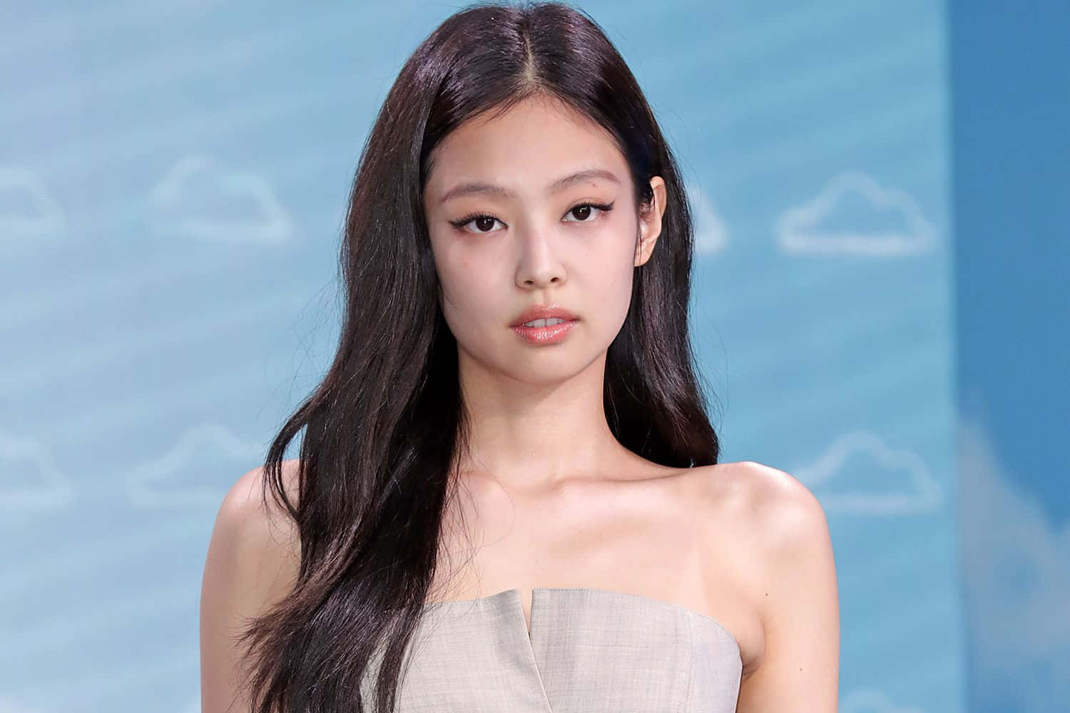 Jennie身穿無肩帶禮服照片 壁紙 — 一位長黑髮的優雅女性，穿著無肩帶米色連衣裙，背景是帶有白雲的柔和藍天。