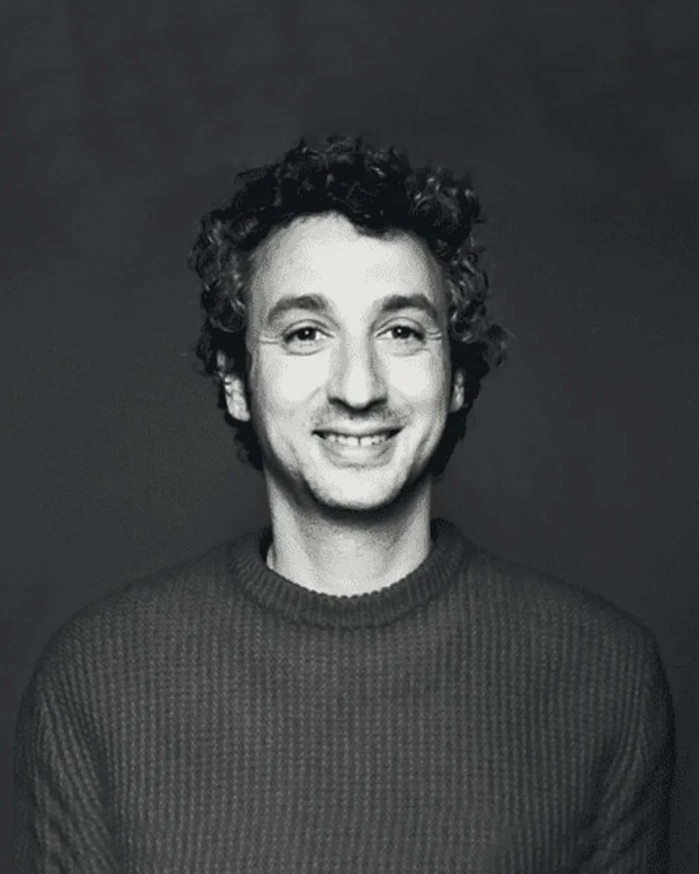 Jérôme Dreyfuss 的灰階肖像 壁紙 — 一個微笑的捲髮男子穿著羅紋毛衣，在深色背景下的黑白肖像。