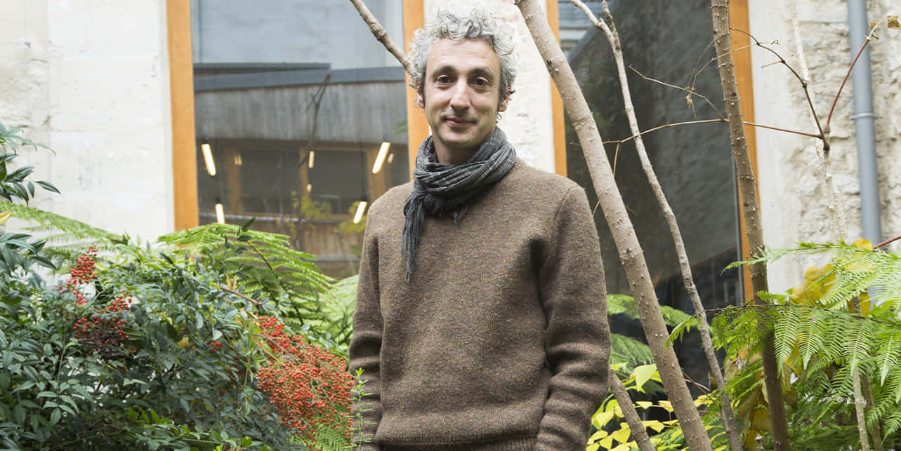 Jérôme Dreyfuss 穿棕色毛衣與灰色圍巾 壁紙 — 一位灰髮男子在花園中微笑，身後有蕨類植物和紅色漿果。