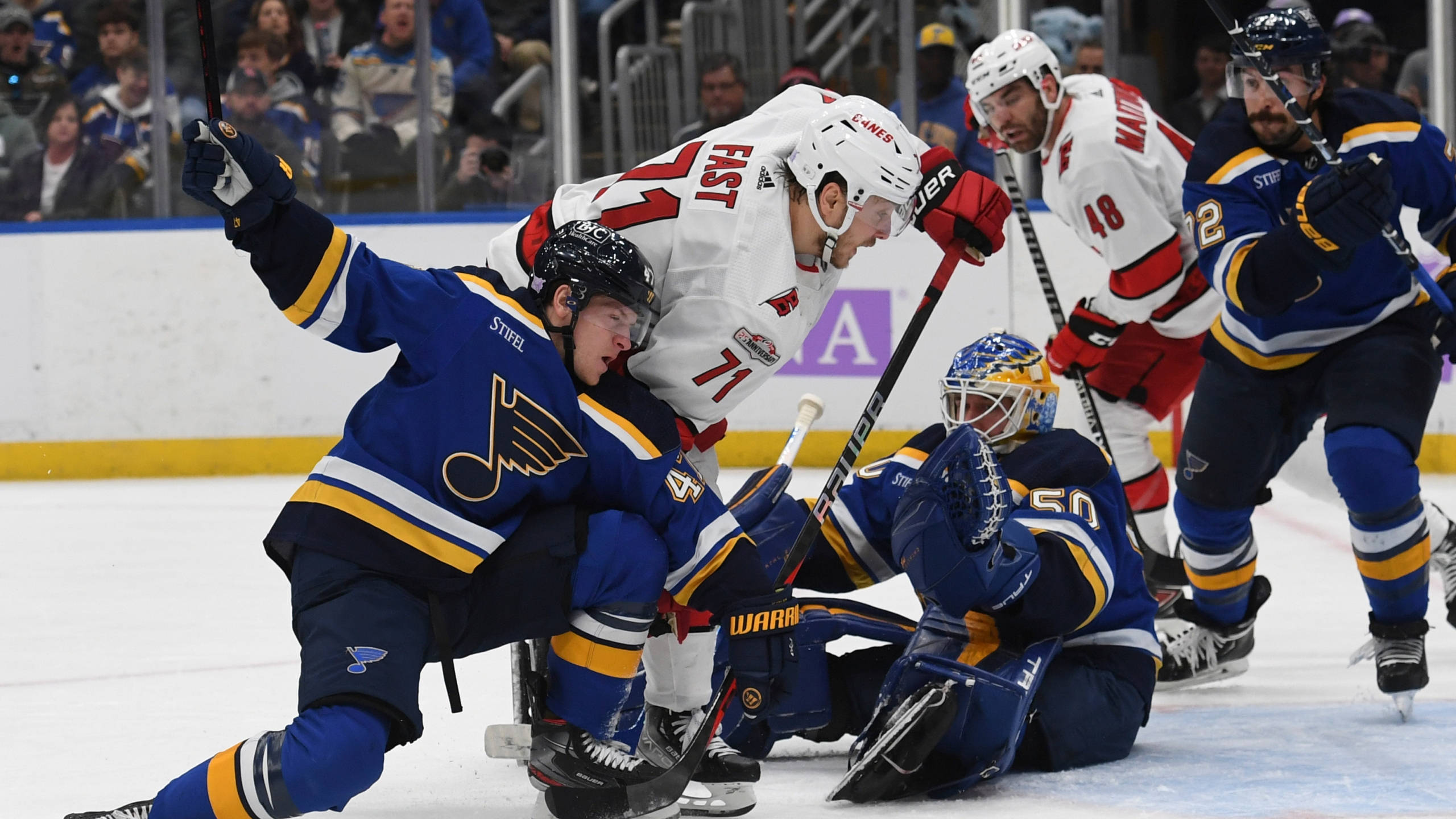Jesper Fast 被 Torey Krug 與 Jordan Binnington 阻擋 壁紙 — 身穿藍白球衣的冰球運動員在快速比賽中圍繞球門激烈爭奪，背景有觀眾。