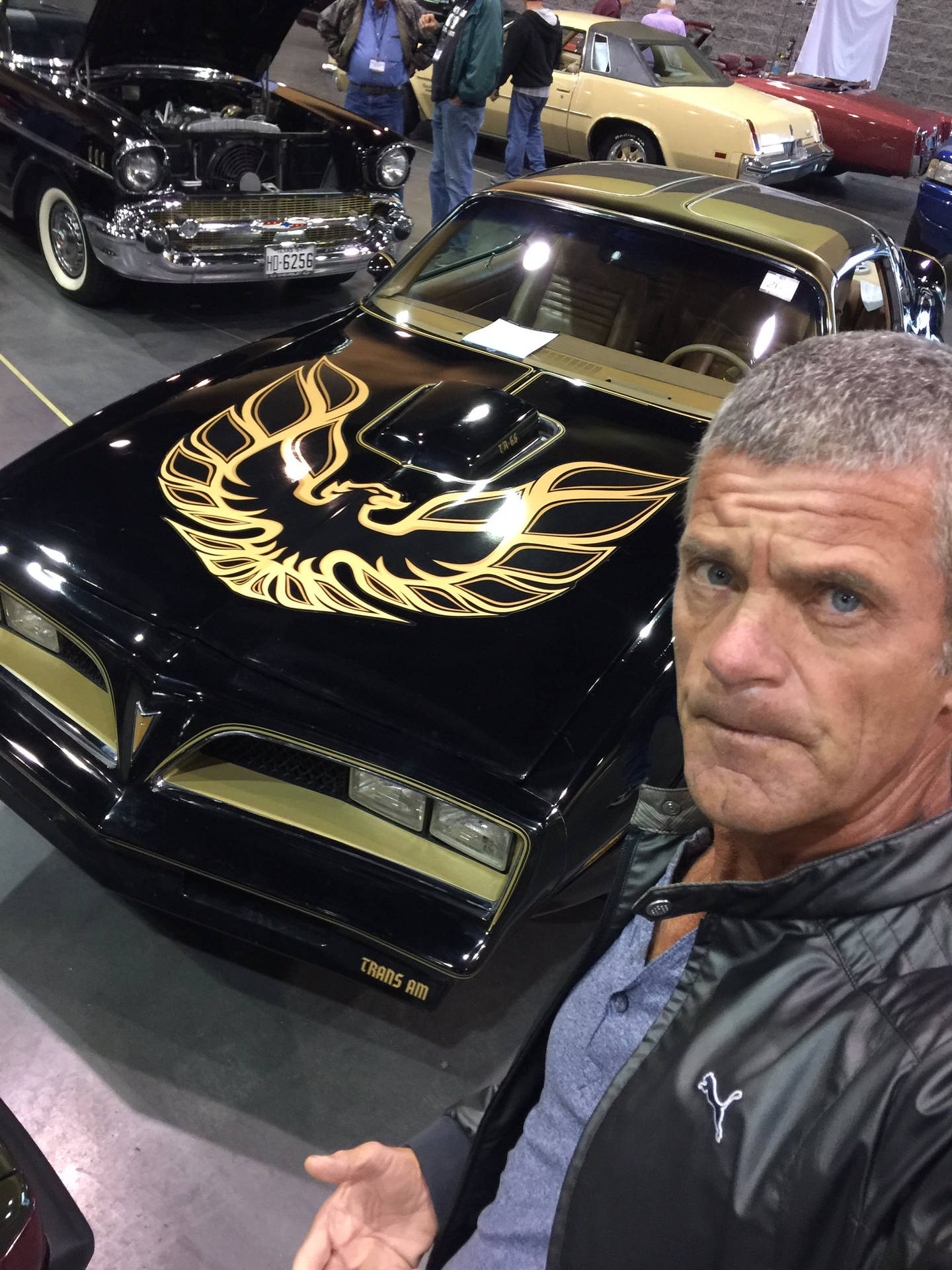 男子站在一輛帶有金色鳳凰貼花的黑色經典汽車旁，在室內車展上。