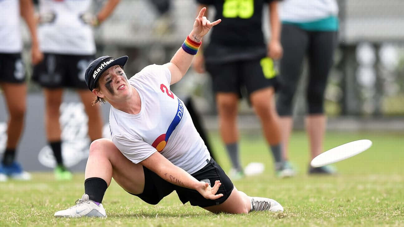 Jesse Shofner 最佳 Ultimate Frisbee 背景 壁紙 — 一名身穿白色球衣和黑色帽子的飛盤高爾夫球手，在投擲帶有彩虹腕帶的飛盤後在草地上滑行。