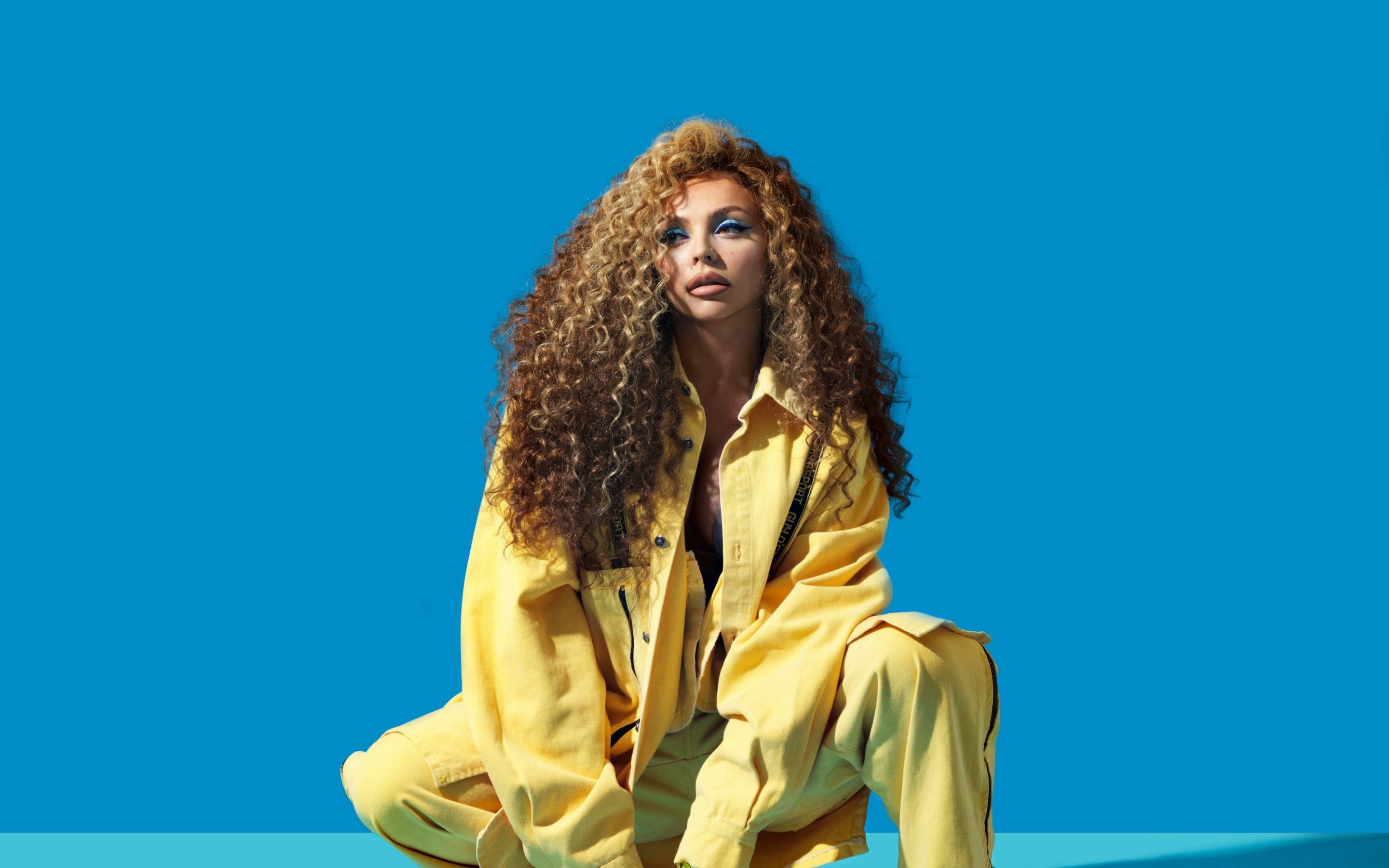 Jesy Nelson 2021 照片拍攝壁紙 壁紙 — 一位擁有豐盈捲髮的女性，身穿亮黃色服裝，坐在鮮豔的藍色背景前。