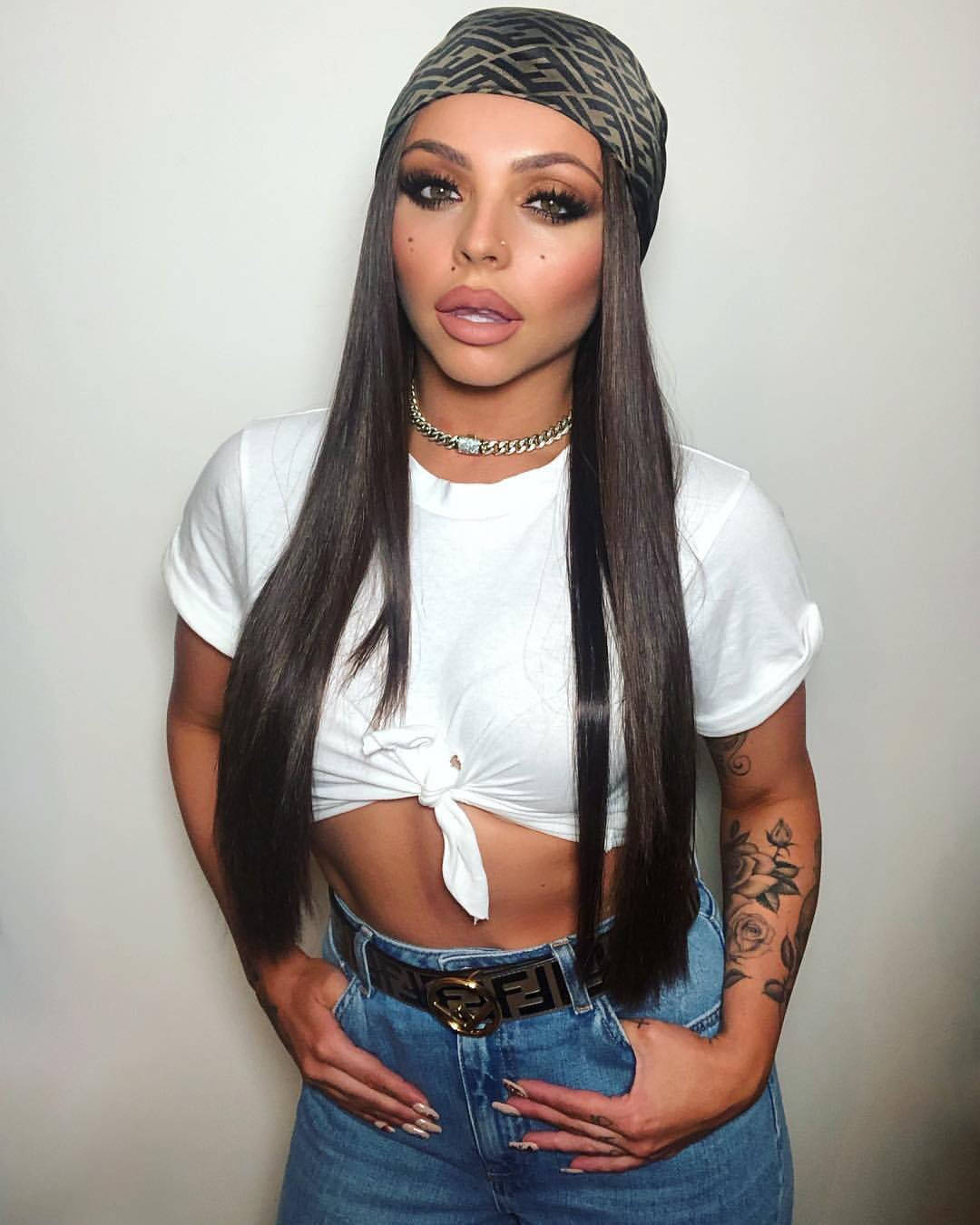Jesy Nelson 潮流酷帥造型 壁紙 — 一位擁有長黑髮和鮮豔妝容的女性，穿著白色綁帶短版上衣和牛仔褲，自信地靠在素牆上擺姿勢。