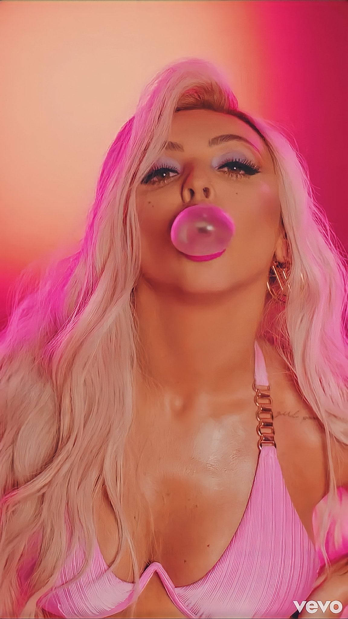 Jesy Nelson 金髮造型 壁紙 — 一位長金髮的女性在粉紅和橙色漸變背景前吹出粉紅色泡泡糖泡泡。