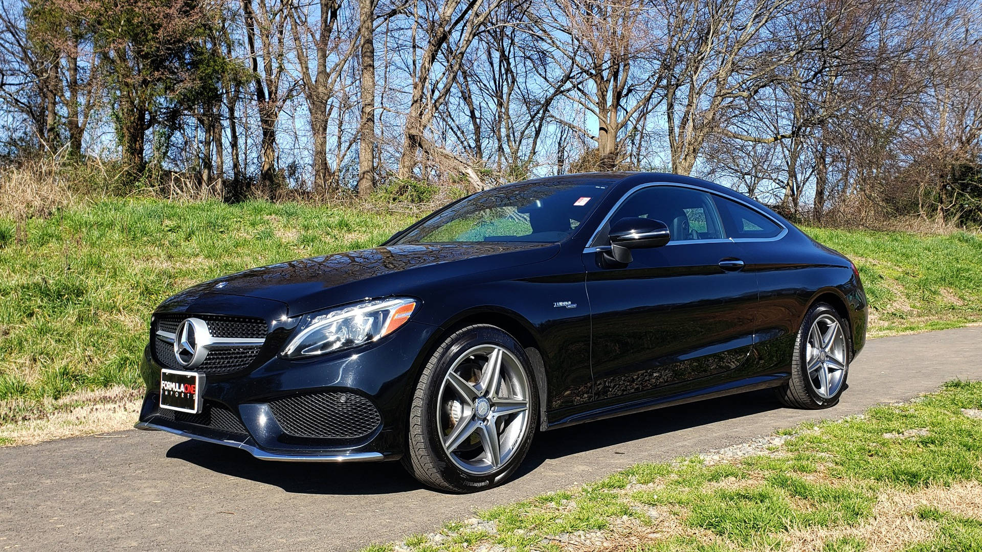 Jet Black 梅賽德斯-奔馳 C300 壁紙 — 一輛時尚的黑色Mercedes C63 AMG雙門轎跑車在明亮陽光下閃耀，停泊在鋪砌道路上，旁邊是草地地形，背景有光禿禿的樹木。