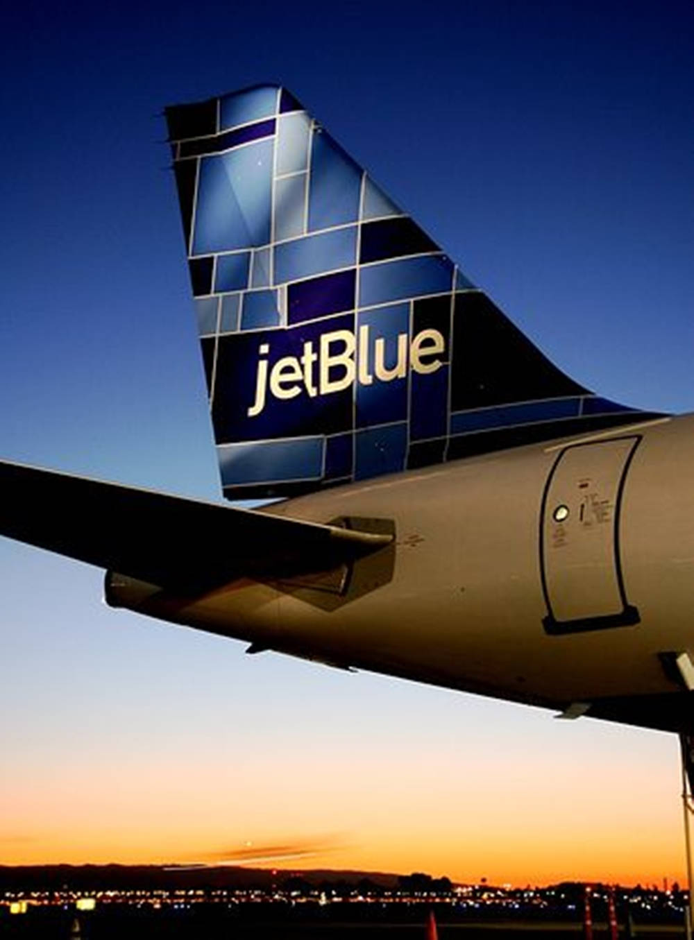 JetBlue航空公司的飛機尾翼印有公司標誌 壁紙 — JetBlue 飛機尾部採用幾何藍色設計，在充滿活力的日落天空下，城市景觀中格外突出。