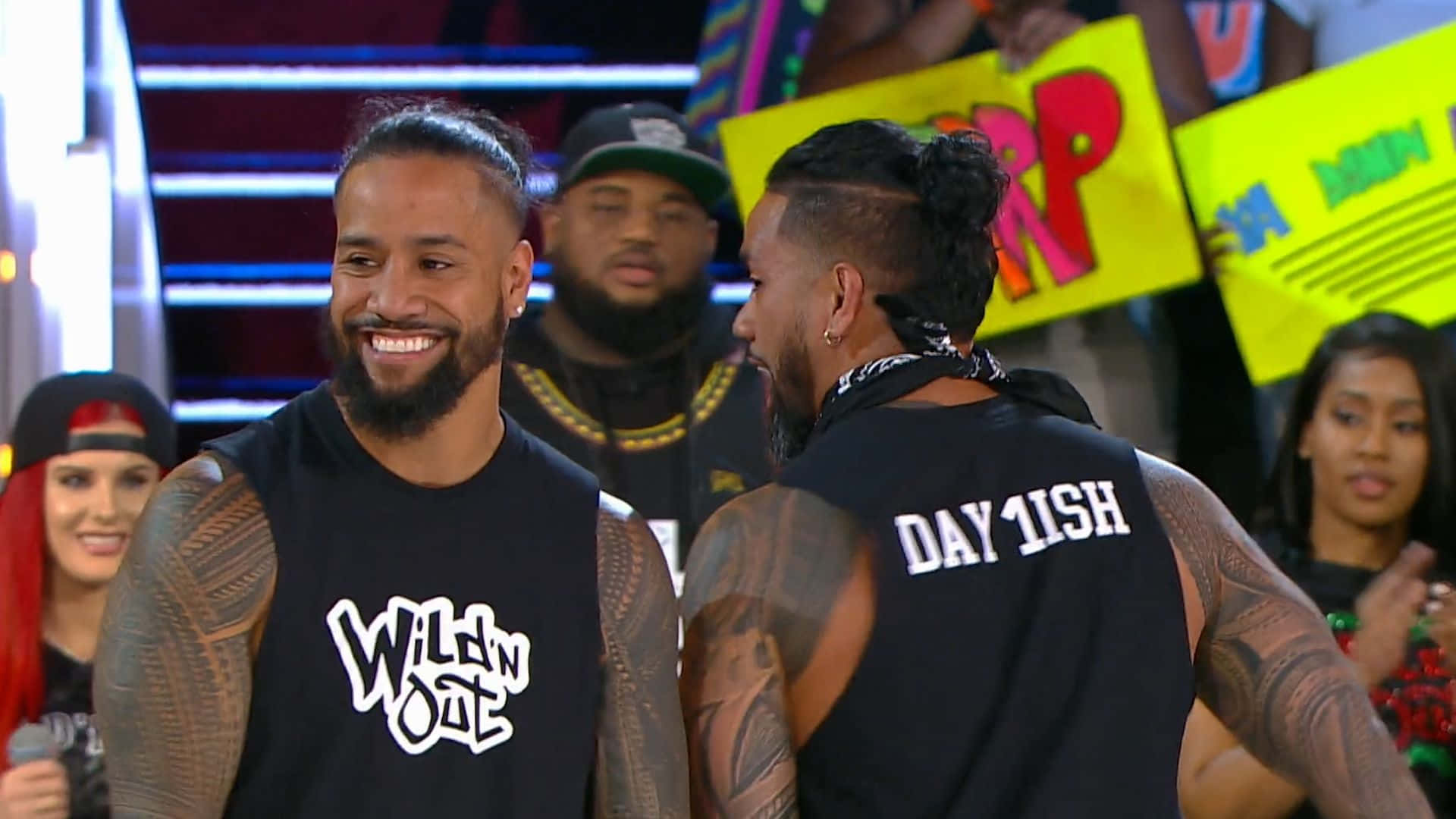 Jey Uso 與 Jimmy Uso 在 WWE 擂台 壁紙 — 兩名紋身男子身穿黑色背心，在燈光璀璨的舞台上微笑互動，身後有粉絲舉著標語牌。