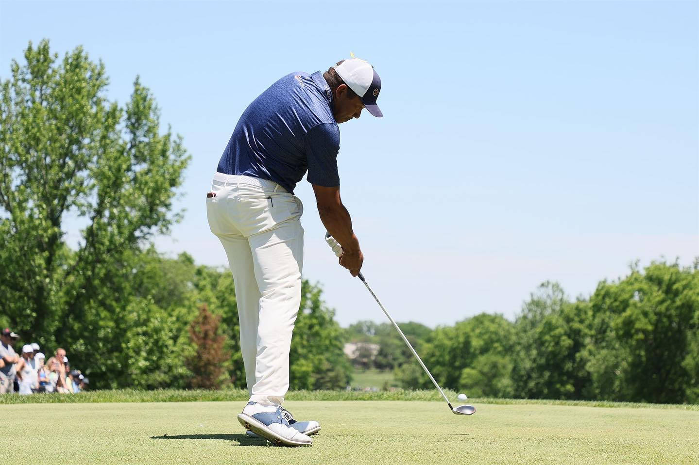 Jhonattan Vegas 後視擊球 壁紙 — 一名身穿藍色襯衫和白色褲子的高爾夫球手在陽光明媚的綠色球場上揮桿，背景有樹木和觀眾。