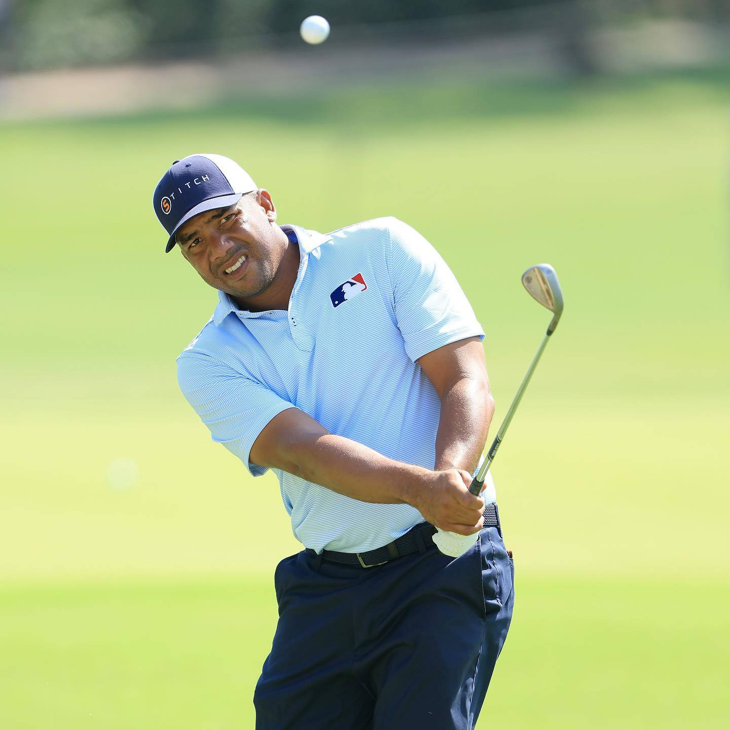 Jhonattan Vegas 注視空中的球 壁紙 — 一名高爾夫球手在陽光明媚的綠色球場上揮桿，高爾夫球在空中，背景模糊。