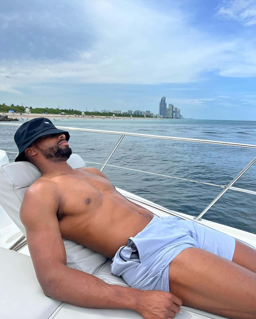 Darius Slayton 桌布 壁紙 — 一名肌肉發達的男子戴著黑色鴨舌帽，赤裸上身躺在白色遊艇甲板上，凝視著部分多雲天空下的沿海城市天際線。