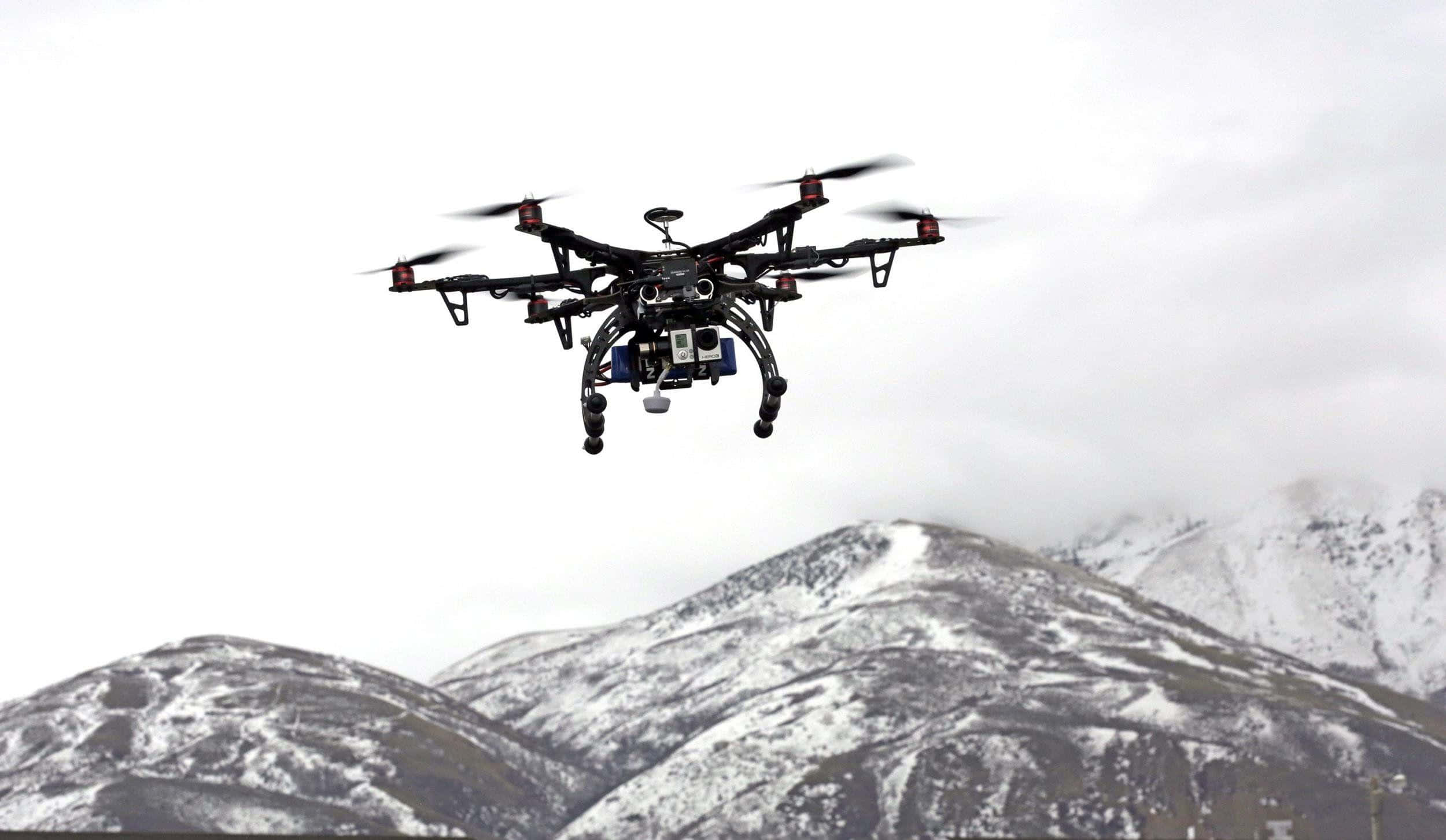 Drone Camera 桌布 壁紙 — 一架黑色六軸無人機在陰天背景下，於積雪山脈前盤旋。