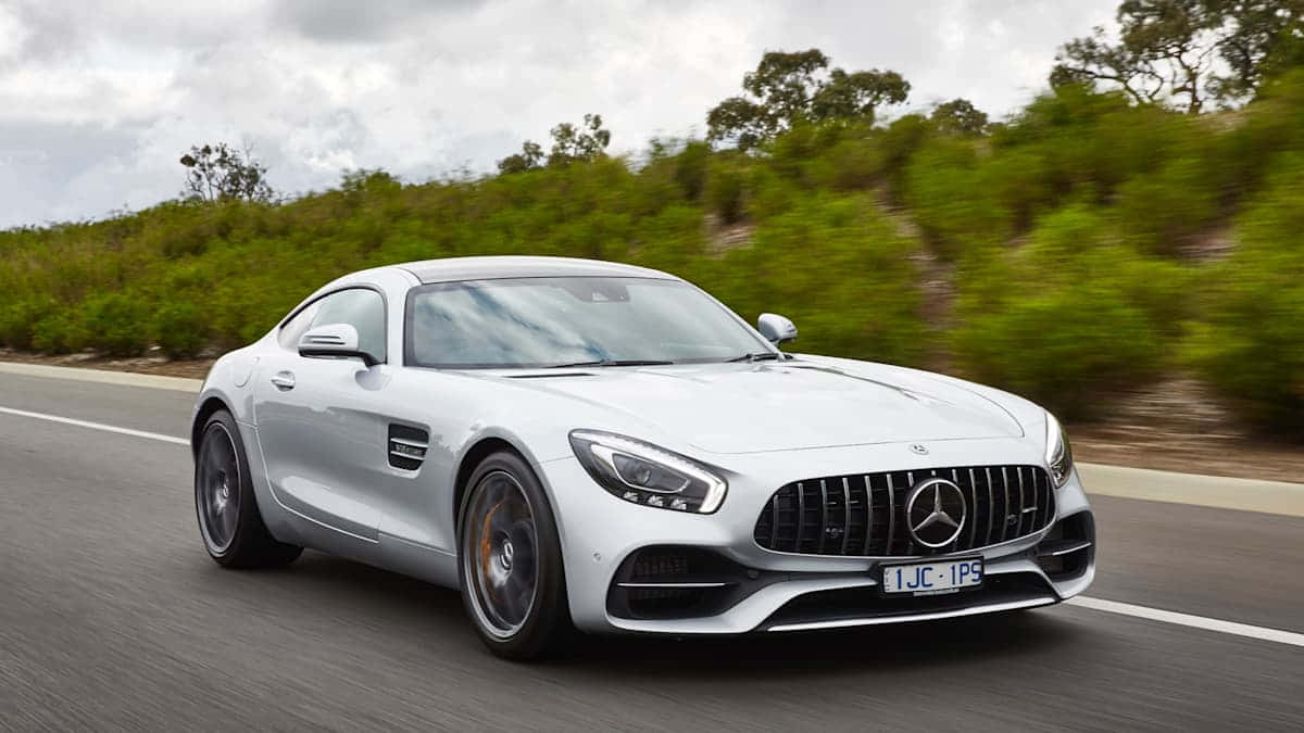 Mercedes Benz Amg Gt 桌布 壁紙 — 一架流線型白色 Mercedes-AMG GT 跑車在陰天之下，沿著蜿蜒的道路穿過翠綠的山丘飛馳。