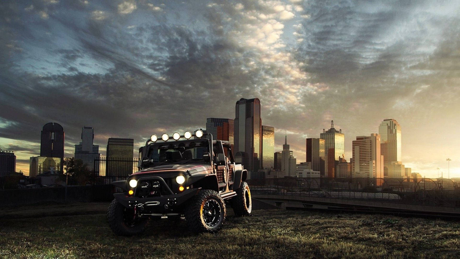 磨砂酒紅色 Wrangler 吉普車 壁紙 — 一輛堅固的黑色越野車，車頭燈閃爍，停在草地上，背景是日落時分高聳的城市天際線。