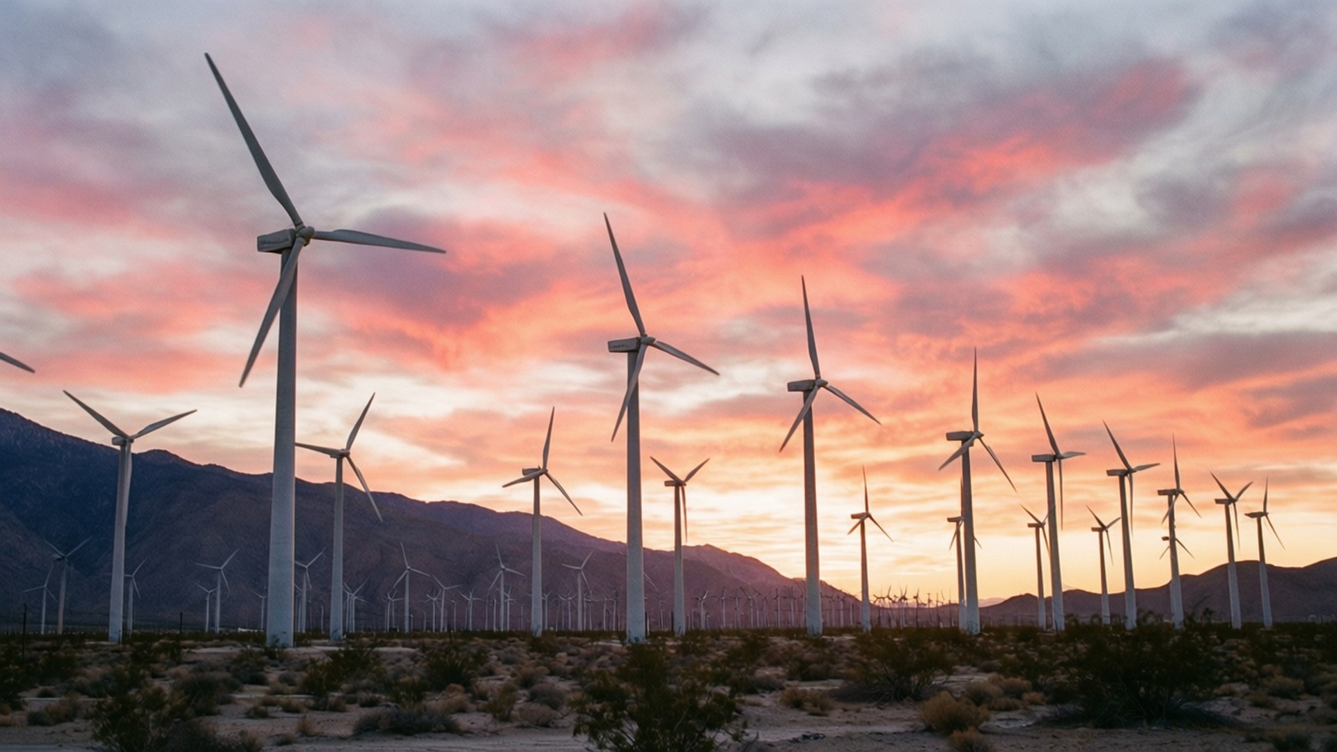 Palm Springs Windmills Sunrise 壁紙 — 剪影風力渦輪機在沙漠日落時分延伸，遠處山巒上方有粉橙色雲彩。