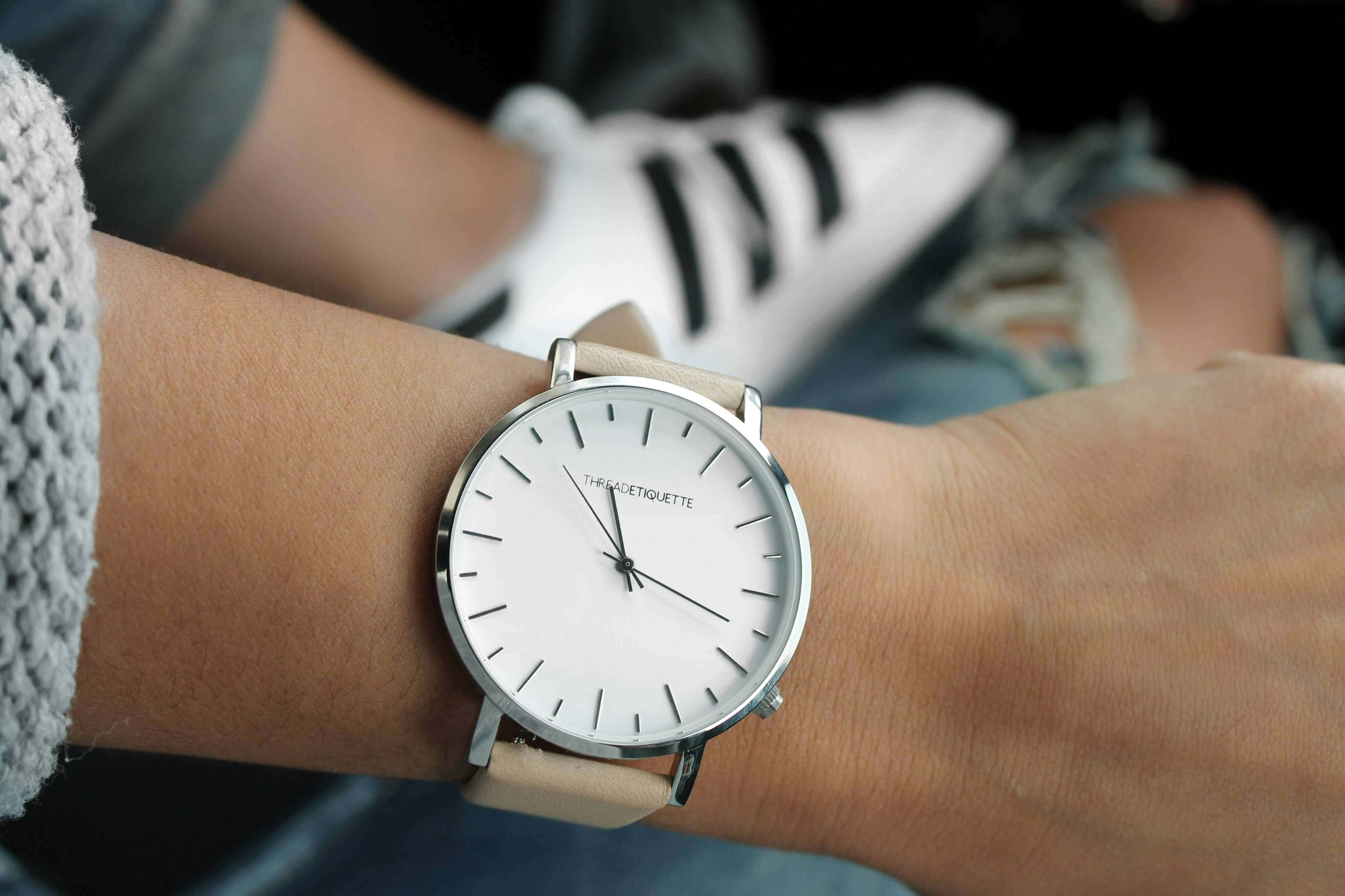Wrist 桌布 壁紙 — 一隻簡約白色手錶配米色皮革錶帶，放在一個人的手腕上，旁邊有一隻白色運動鞋和灰色針織袖。