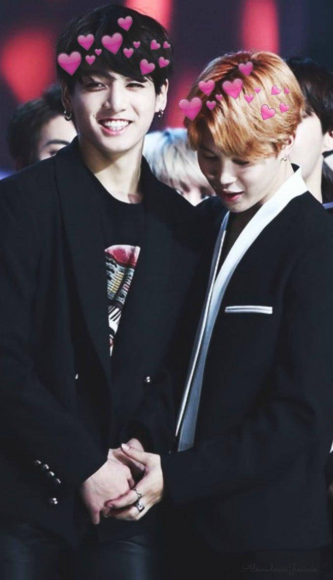 Jikook 手拉手戴心形髮飾 壁紙 — 兩個微笑的黑西裝男人牽手，粉紅色心形圖案漂浮在他們頭頂，背景是模糊的紅色。