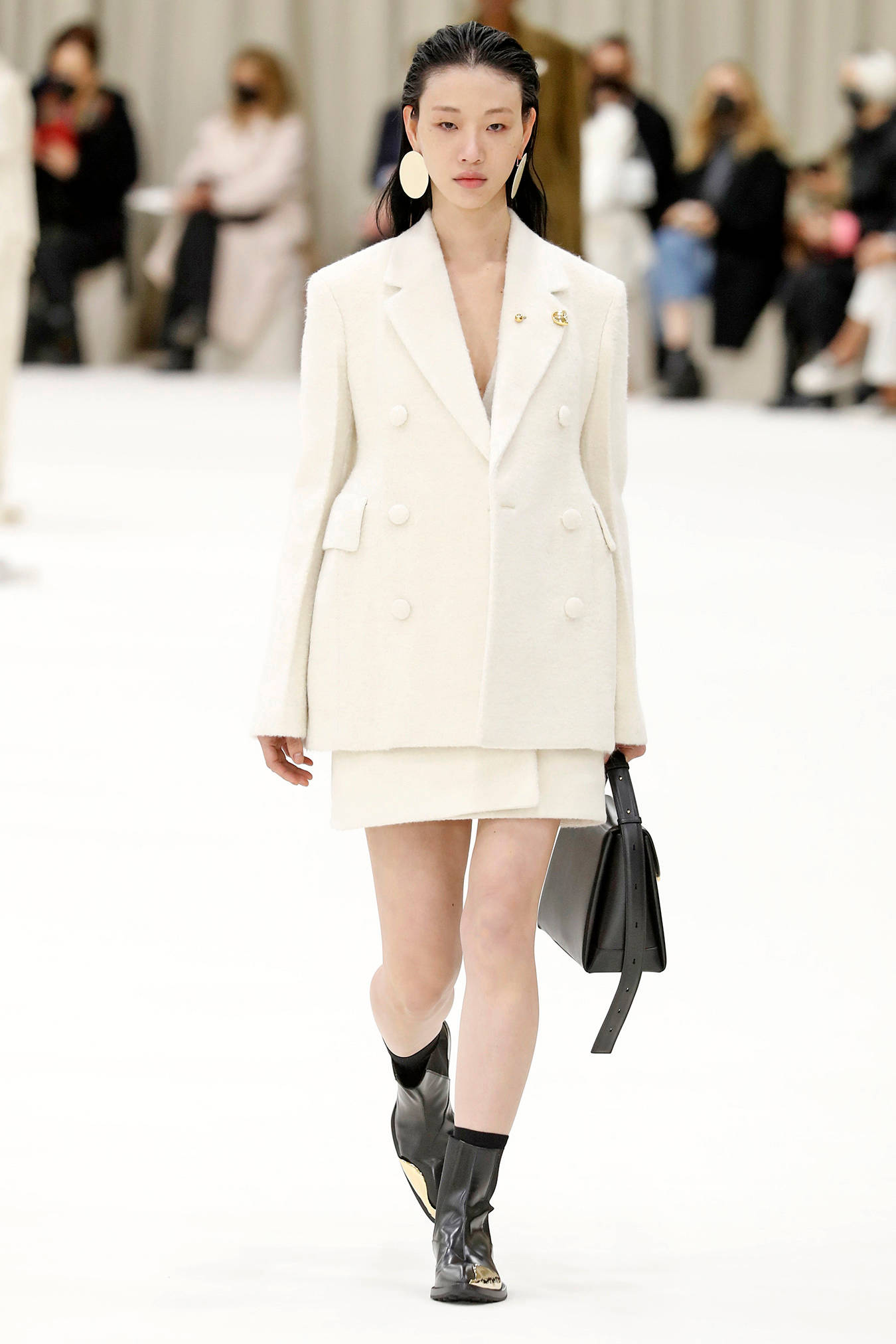 Jil Sander 精緻白色禮服西裝 壁紙 — 一位模特兒自信地走在天橋上，身穿時尚的白色雙排扣西裝外套和配套裙子，搭配黑色靴子和結構感黑色手袋。