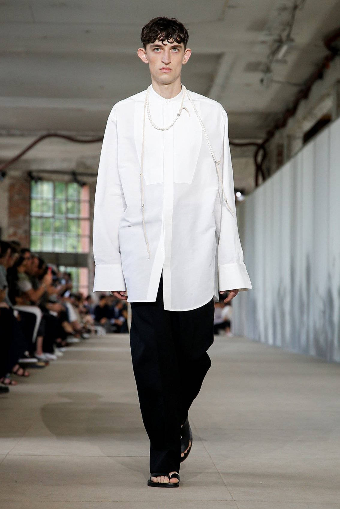 Jil Sander 黑褲 壁紙 — 一位男模特兒在明亮燈光下，穿著簡約白色寬鬆襯衫和黑色長褲，在混凝土天橋上行走。