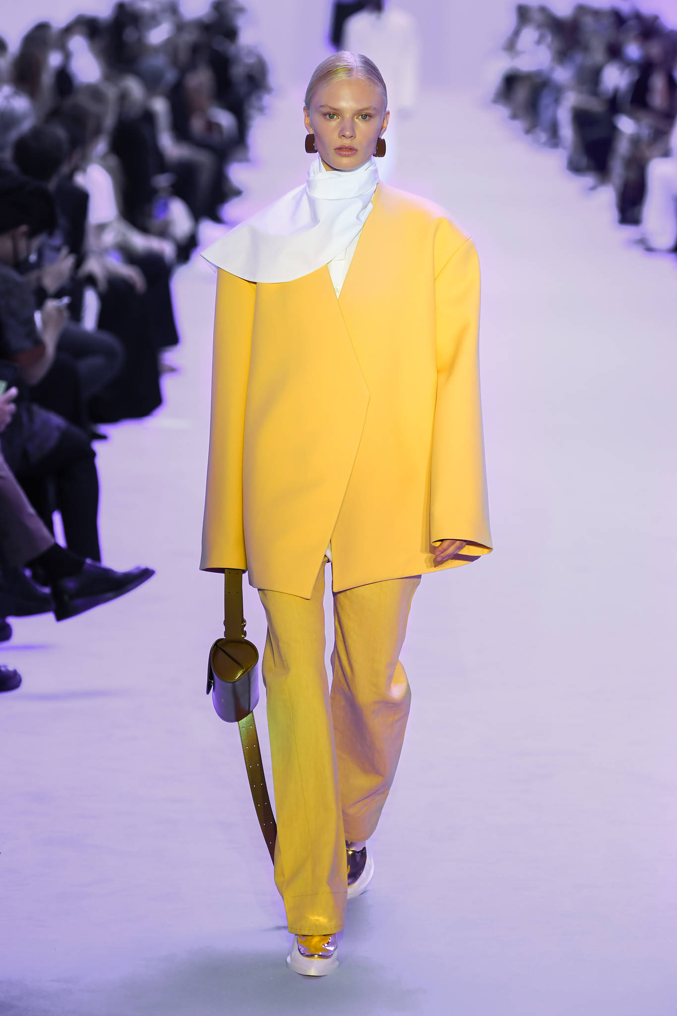 Jil Sander 黃色長袖連衣裙 壁紙 — 一位模特兒穿著大膽的黃色西裝，白色領口，拿著棕色手袋，走在天橋上。