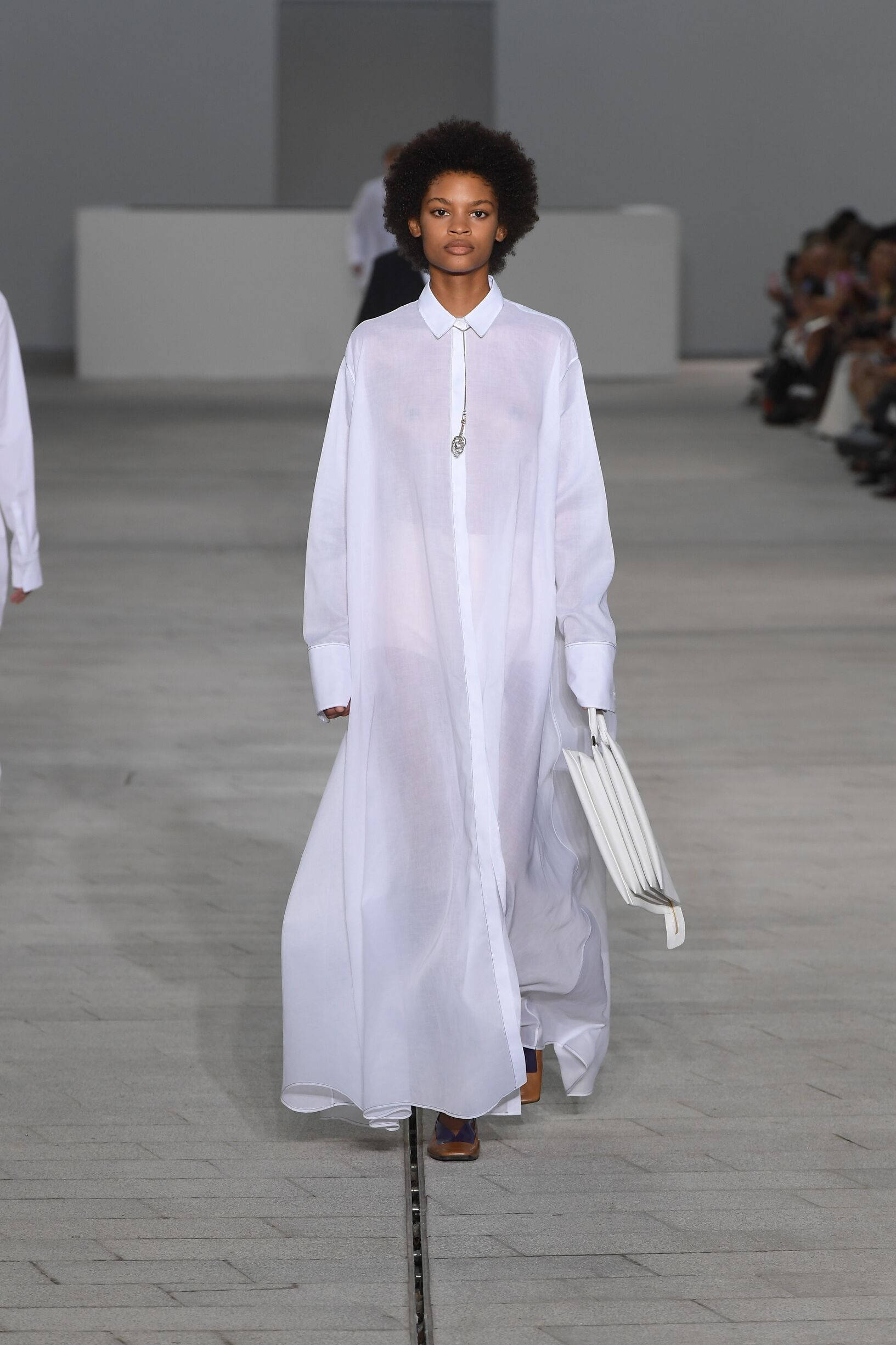 Jil Sander 白色長袖 壁紙 — 一位擁有非洲爆炸頭髮型的模特兒，自信地走在簡約的T台上，身穿飄逸的白色襯衫裙，手持白色結構感手袋。