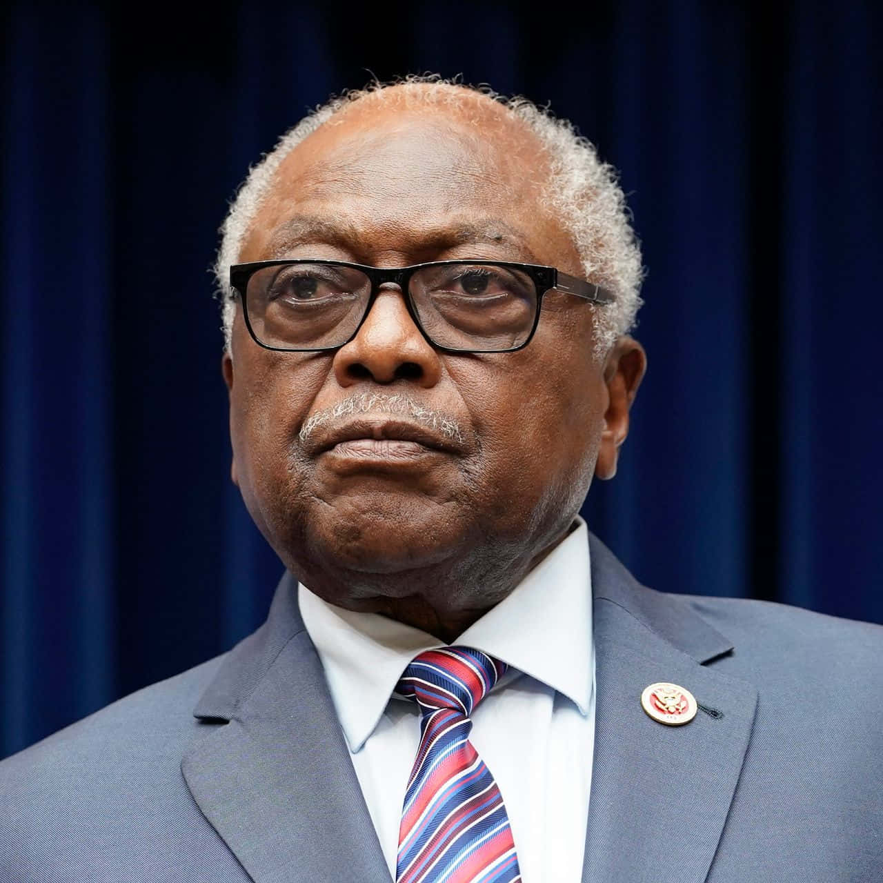Jim Clyburn 面露嚴肅 壁紙 — 一位戴眼鏡、打條紋領帶、表情嚴肅的年長黑人男子，身穿正裝站在深藍色窗簾前。