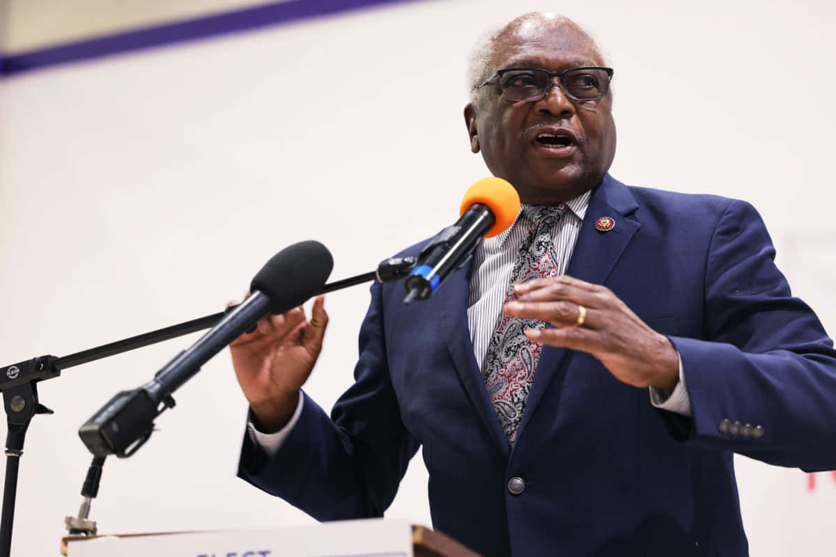 美國眾議員 Jim Clyburn 與媒體對話 壁紙 — 一位年長的黑人男子身穿藍色西裝和圖案領帶，雙手比劃著，熱情地對著麥克風說話。