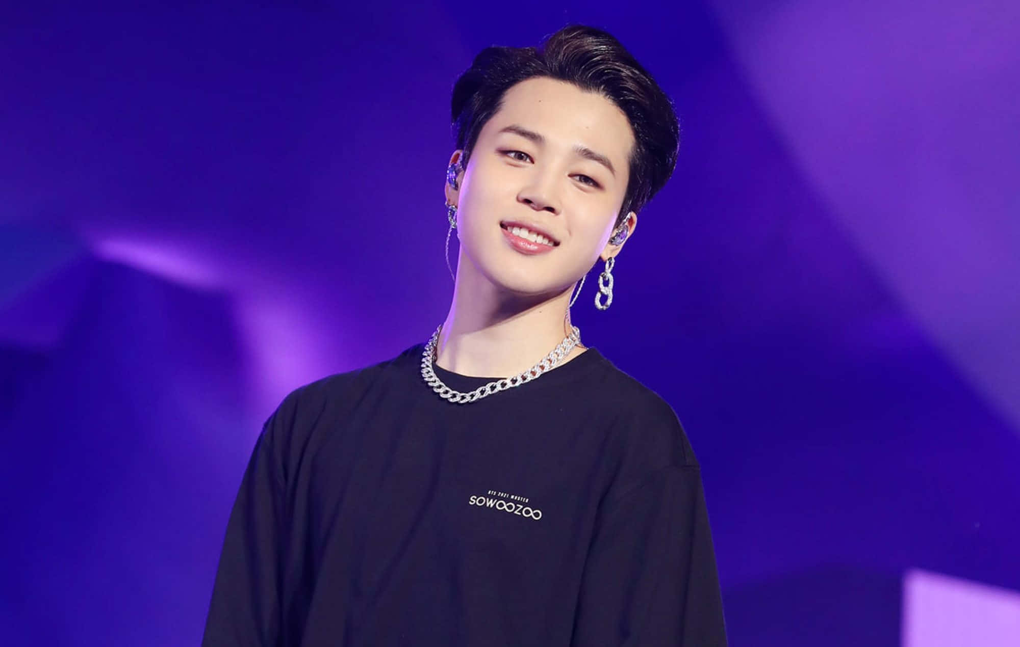 Jimin BTS 以燦爛笑容俘獲人心 壁紙 — 一位微笑的年輕男子，頭髮造型時尚，戴著銀色耳環，身穿黑色T恤，背景是充滿活力的紫色舞台。