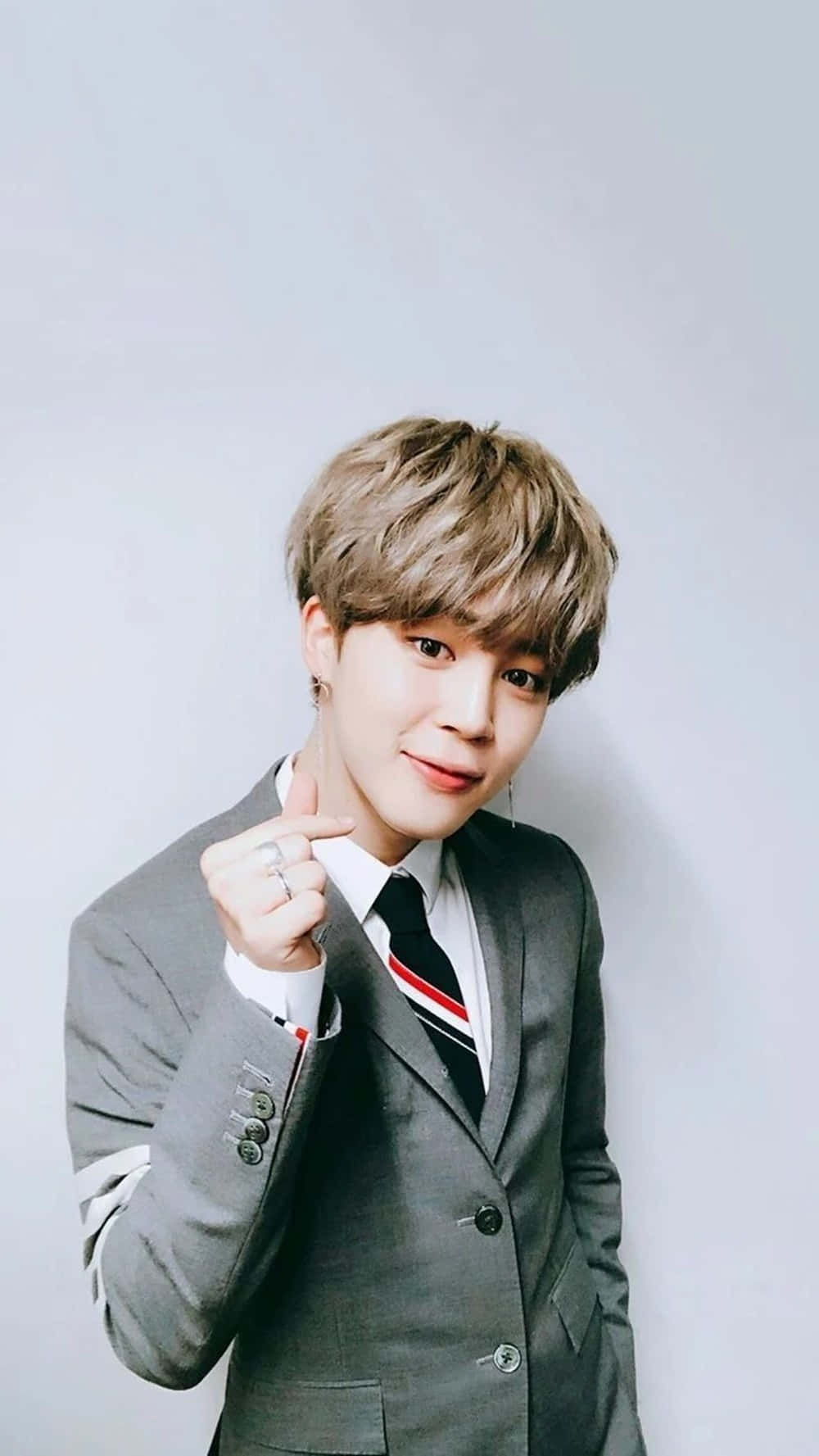 Jimin BTS 指心灰色西裝 壁紙 — 一位身穿灰色西裝、打著條紋領帶的年輕男子，在純淺灰色背景前微笑著指向觀看者。