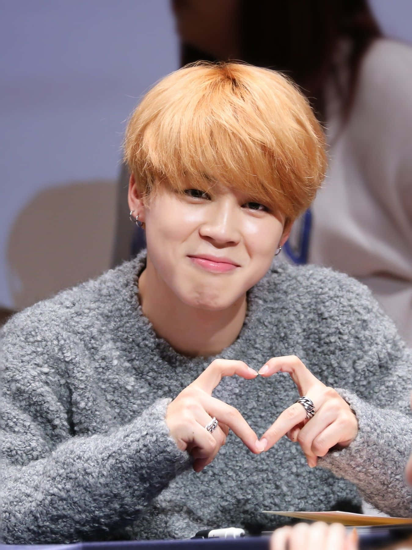 Jimin BTS 指心粉絲見面會 壁紙 — 一位橙髮年輕男子戴著戒指，雙手比出心形，溫暖地微笑。