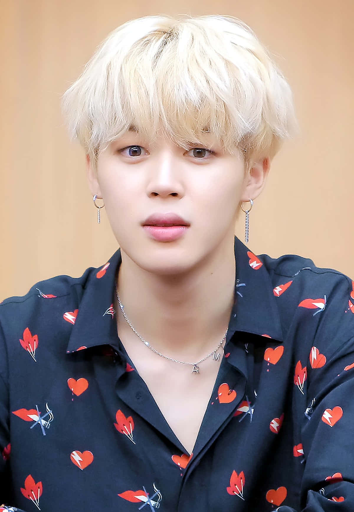 Jimin 主導舞台，以迷人舞姿吸引目光 壁紙 — 一位年輕男子，擁有鉑金色頭髮和心形圖案襯衫，在溫暖的米色背景前溫柔地凝視鏡頭。