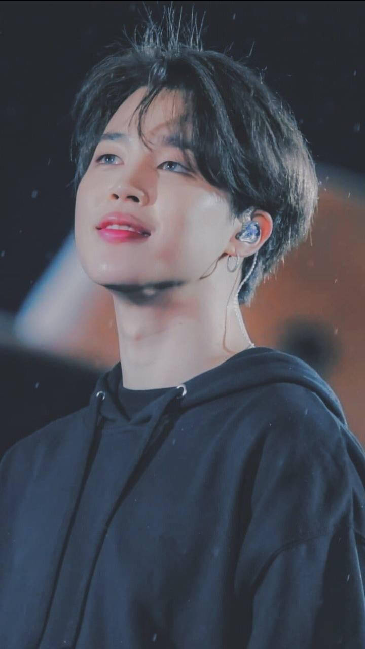 Jimin 美學仰拍視角 壁紙 — 一位黑髮紅唇的年輕男子在舞台燈光下穿著黑色連帽衫和耳機，向上凝視。