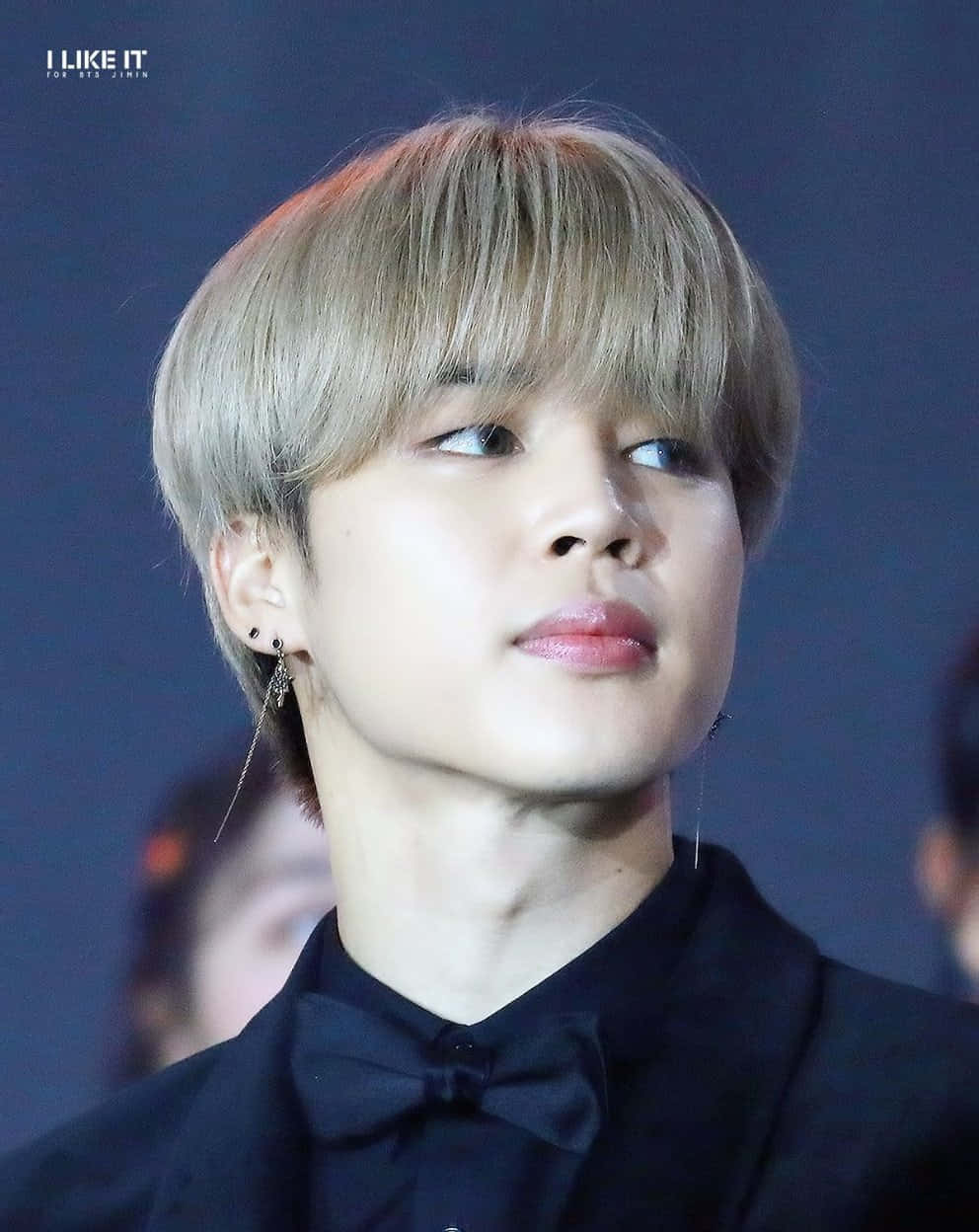 Jimin 舞台光芒四射 壁紙 — 一個年輕金髮男子戴著黑色蝴蝶結領結，在深藍色背景下向上看的特寫肖像。