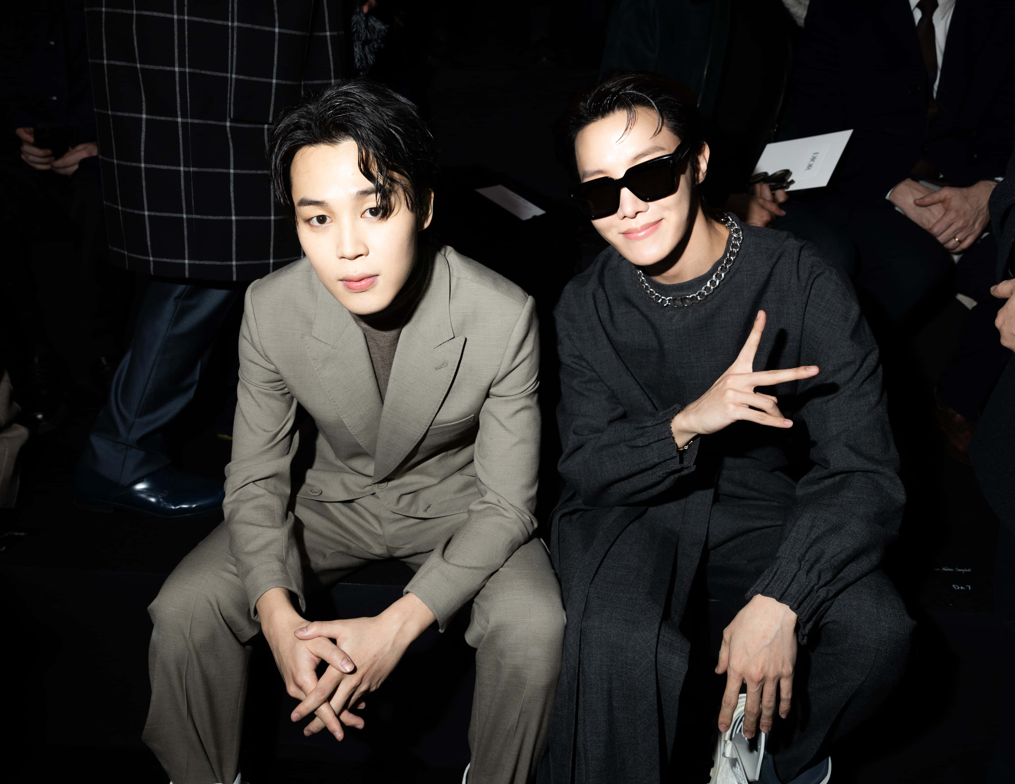 Jimin慶祝巨大成功 壁紙 — 兩位時尚男士坐在時裝秀上，一位身穿米色西裝，另一位身穿黑色服裝戴太陽眼鏡比出和平手勢。