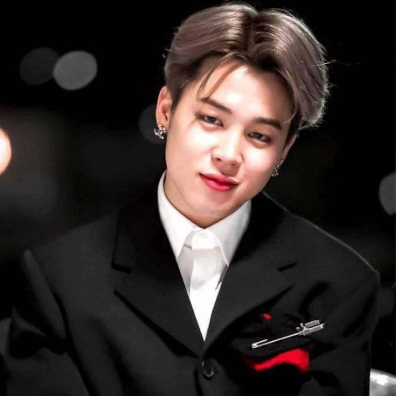 Jimin望向未來，充滿希望 壁紙 — 一位年輕男子，梳著時尚髮型，戴著耳環，身穿黑色西裝，口袋裡放著紅色方巾，背景是模糊的深色。