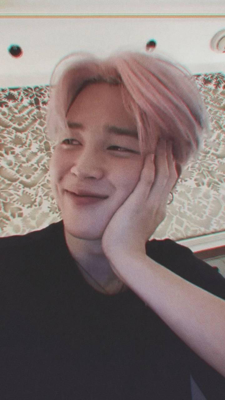 Jimin美學燦爛微笑 壁紙 — 一位粉紅色頭髮的年輕男子微笑著，將臉頰靠在手上，背景是圖案壁紙。
