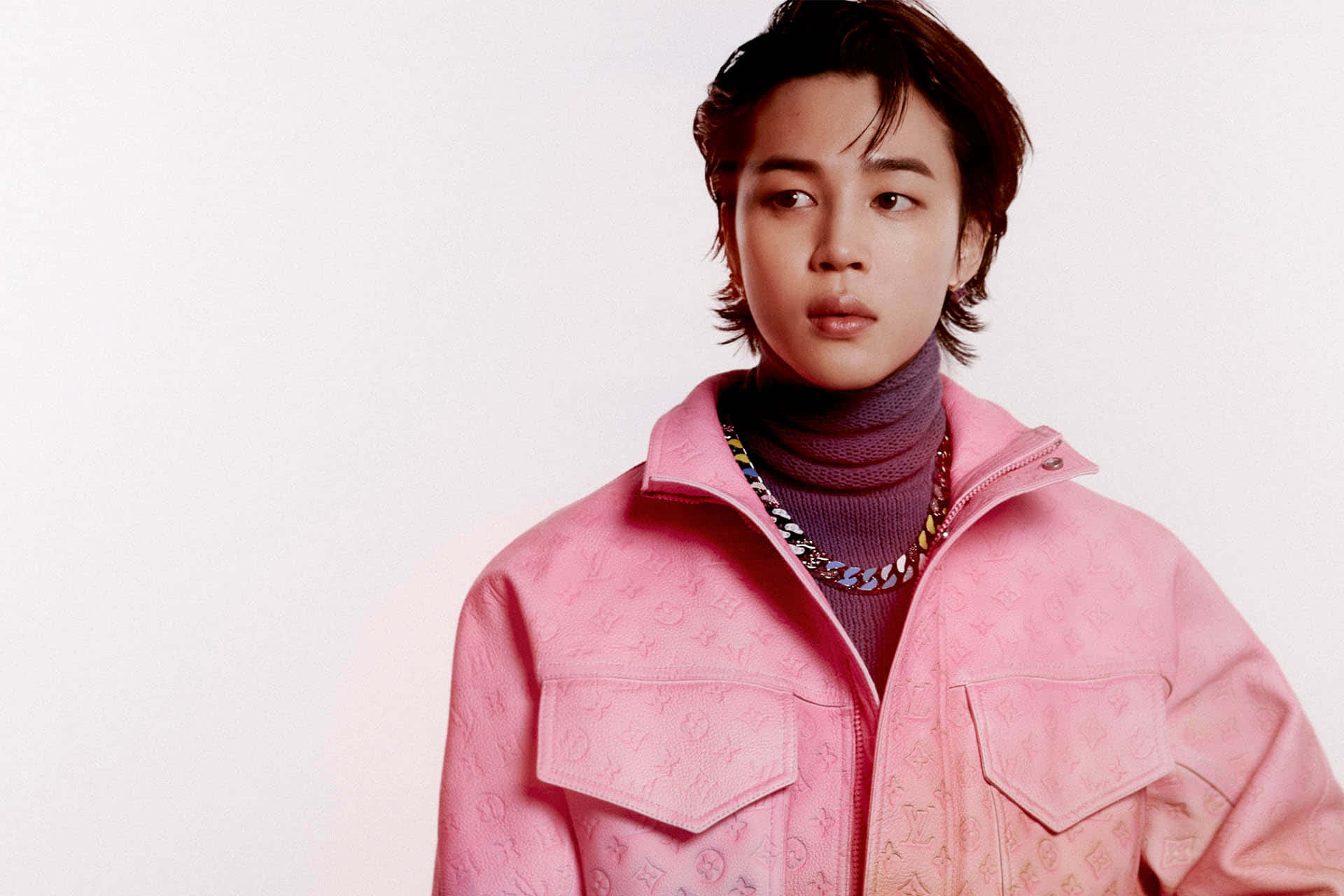 Jimin身著經典黑色西裝站在聚光燈下 壁紙 — 一名身穿粉紅色字母圖案外套和紫色高領毛衣的年輕男子，在純白背景前若有所思地凝視。