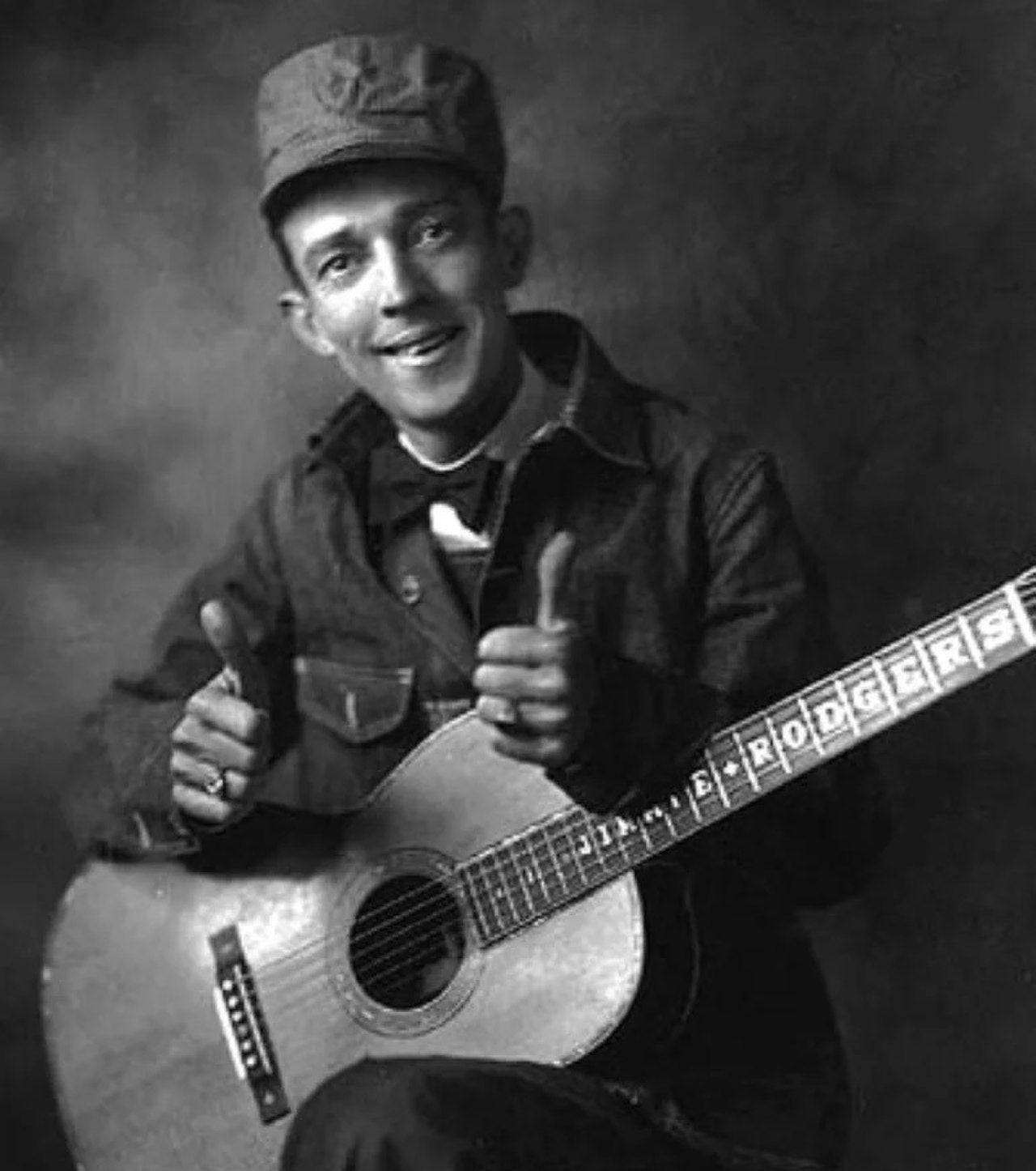 Jimmie Rodgers 黑白肖像照 壁紙 — 一個戴帽微笑的男人豎起兩個大拇指，同時抱著一把指板上有他名字的木結他。