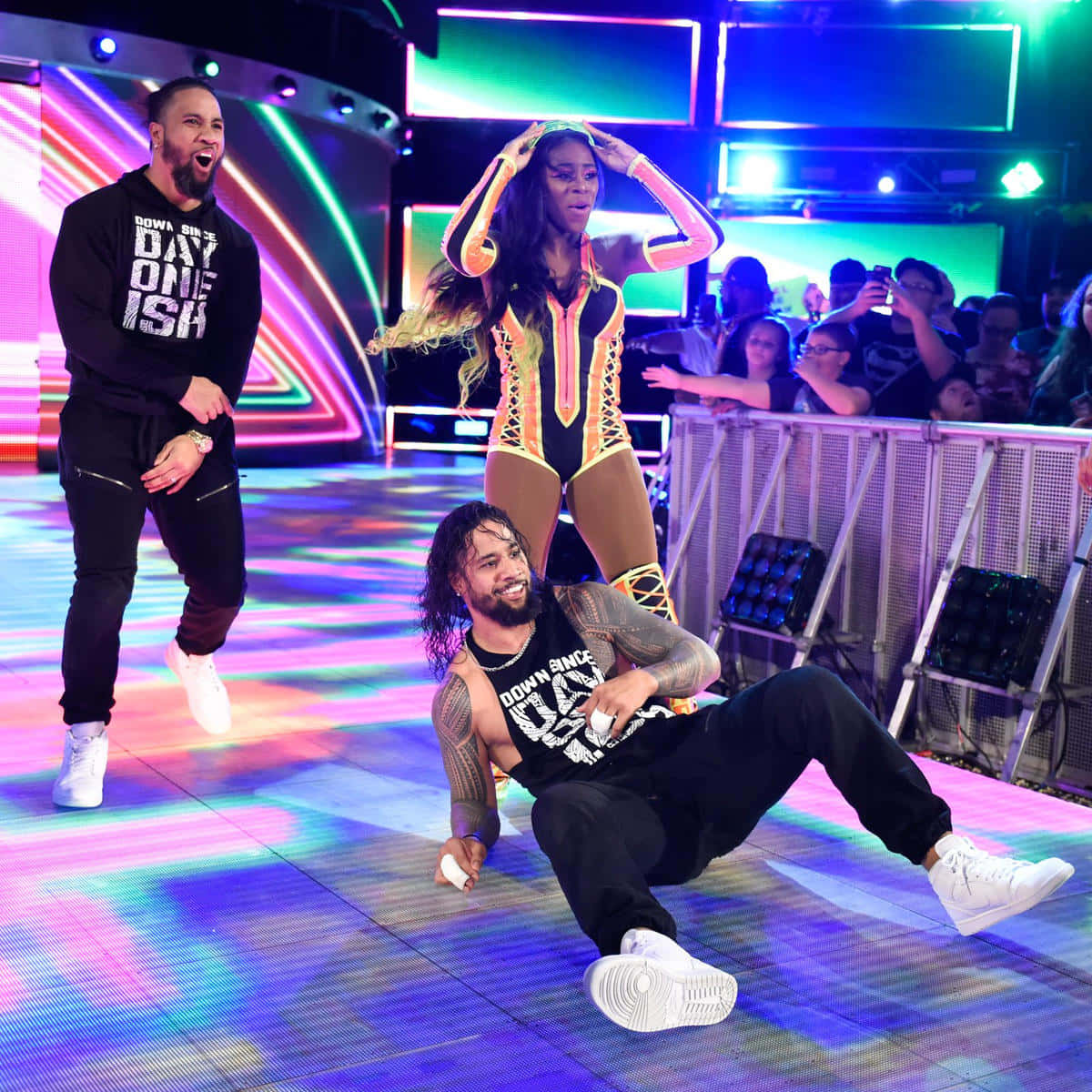 Jimmy 與 Jey Uso 與 WWE 花樣女郎 Naomi 同框 壁紙 — 三名摔角手在充滿活力的霓虹燈舞台上精力充沛地表演，背後有歡呼的人群隔著屏障。