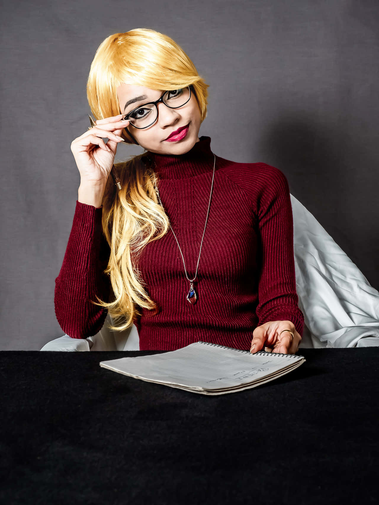 Harleen Quinzel 桌布 壁紙 — 一位金髮戴眼鏡的女性坐在一張有筆記本的暗色桌子旁調整眼鏡。