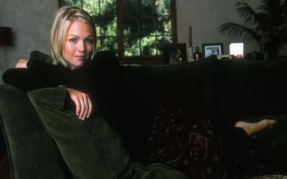Jennie Garth 桌布 壁紙 — 一位金髮女郎在昏暗舒適的客廳裡，放鬆地躺在深綠色天鵝絨沙發上，窗外可見樹木。